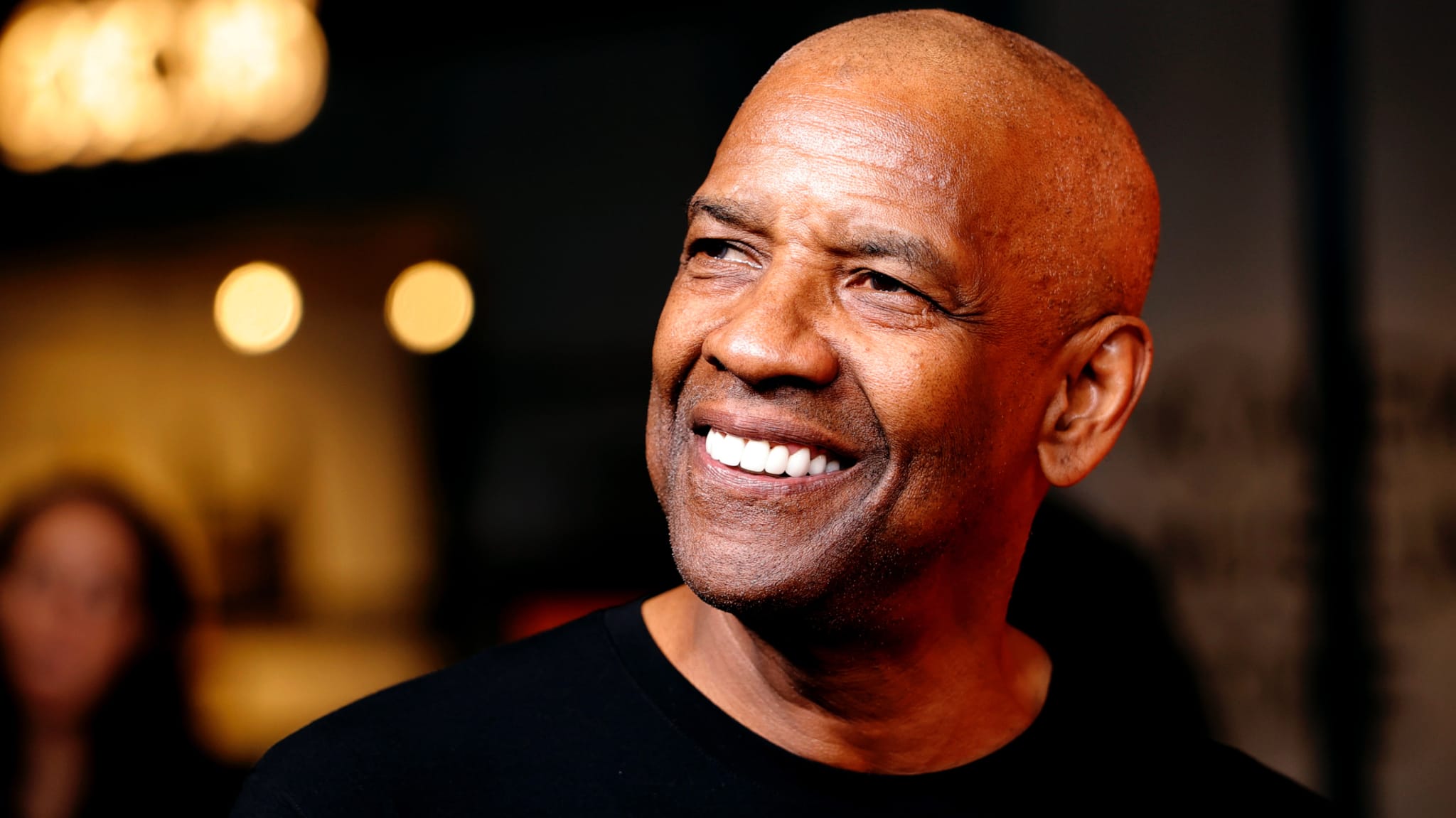 Denzel Washington smiling, wearing a black shirt, with blurred lights in the background.