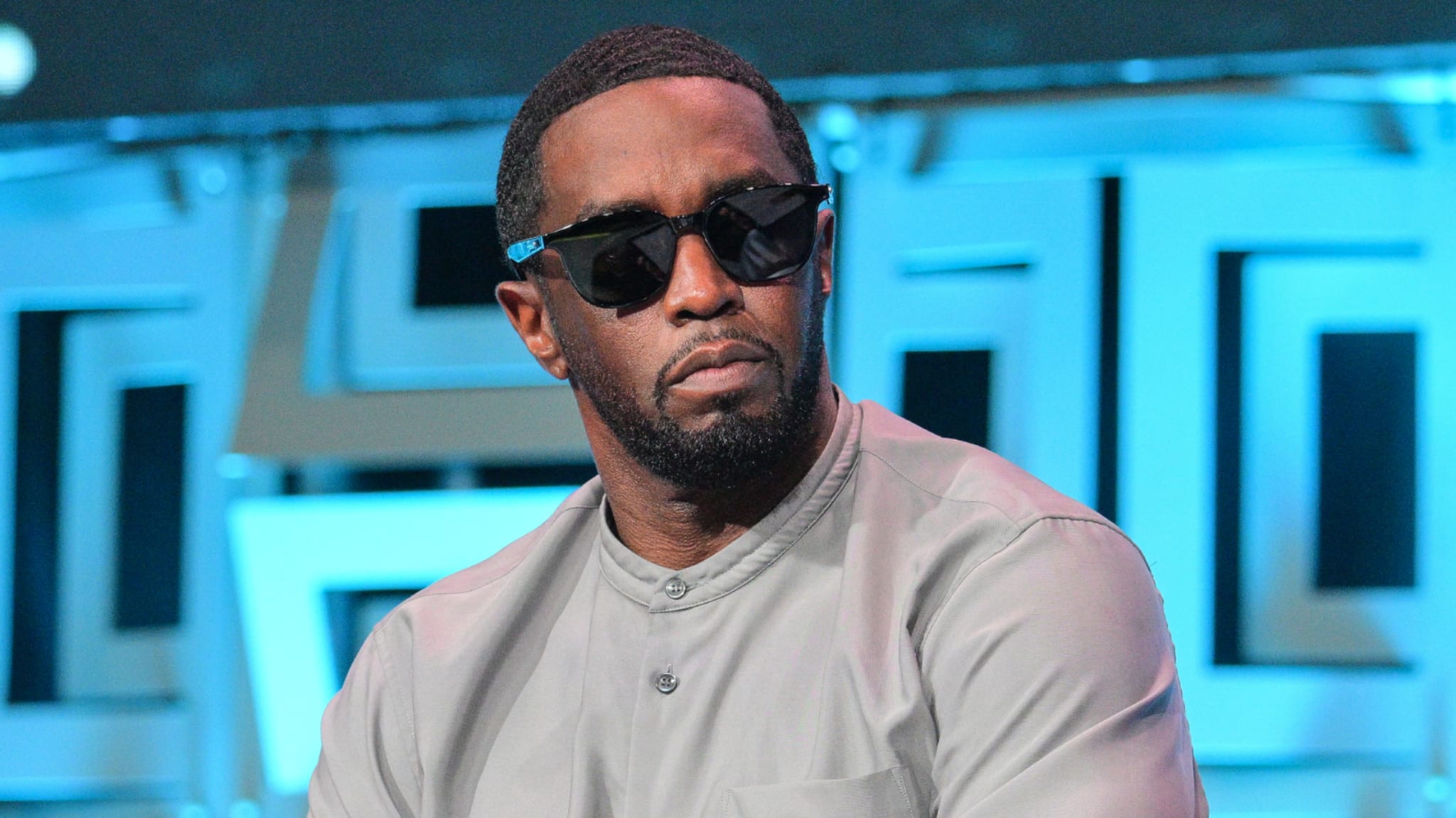 Sean "Diddy" Combs wearing sunglasses and a light gray shirt, sitting against a blue geometric background.