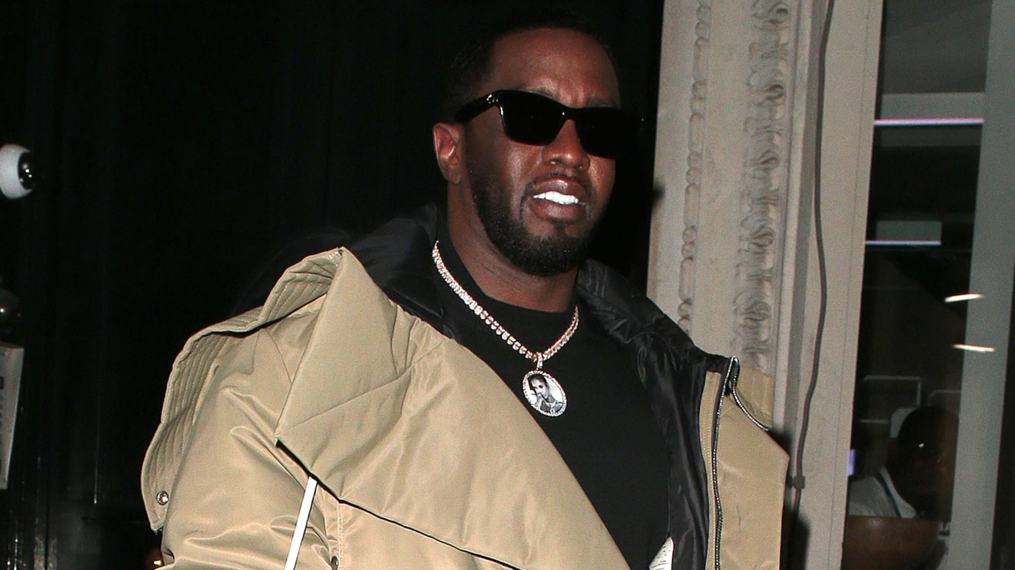 Sean "Diddy" Combs wearing sunglasses and a beige coat, accessorized with a necklace, walking outside at night.