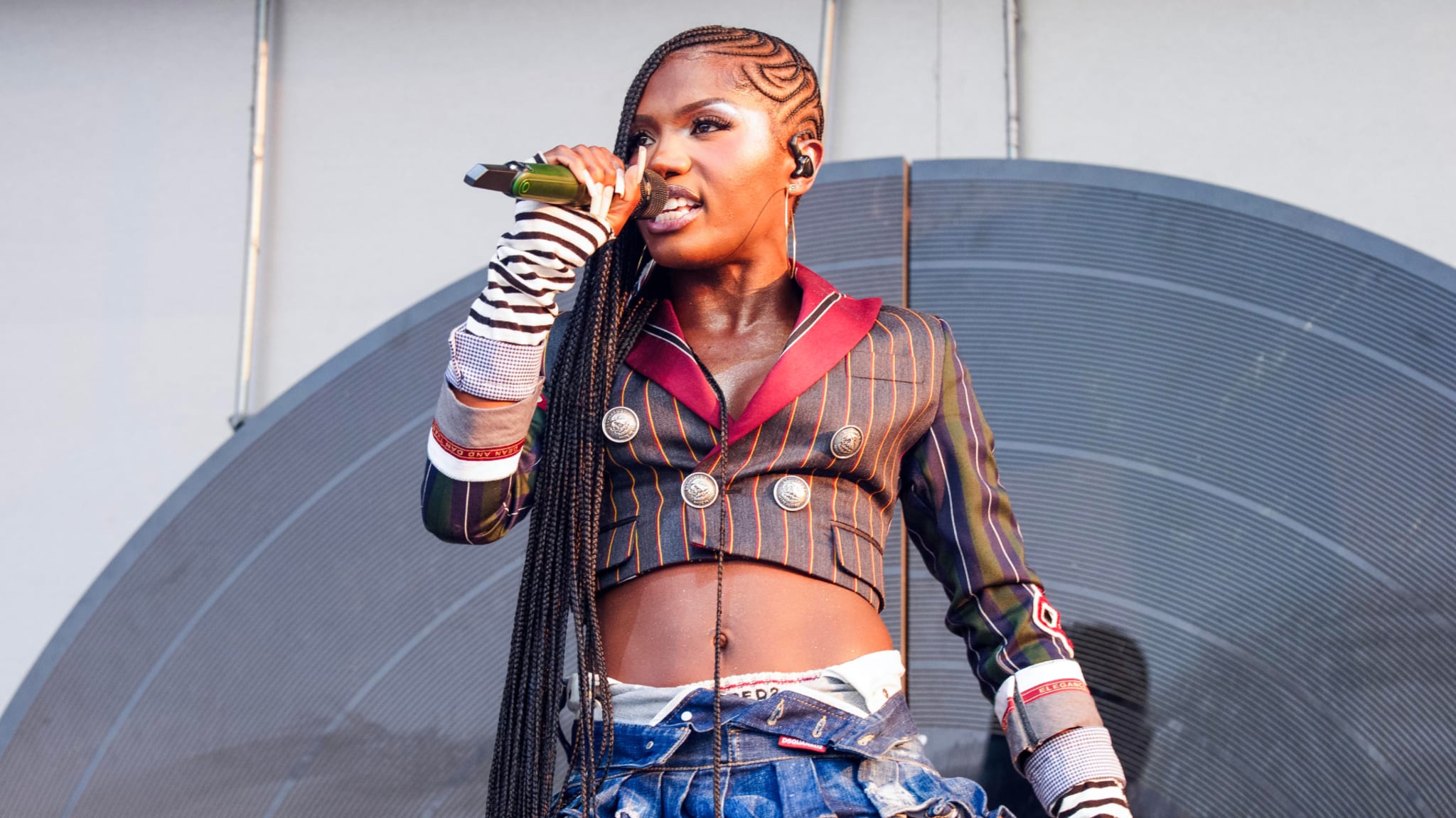 Doechii on stage with long braids, wearing a stylish cropped jacket and denim pants, holding a microphone.