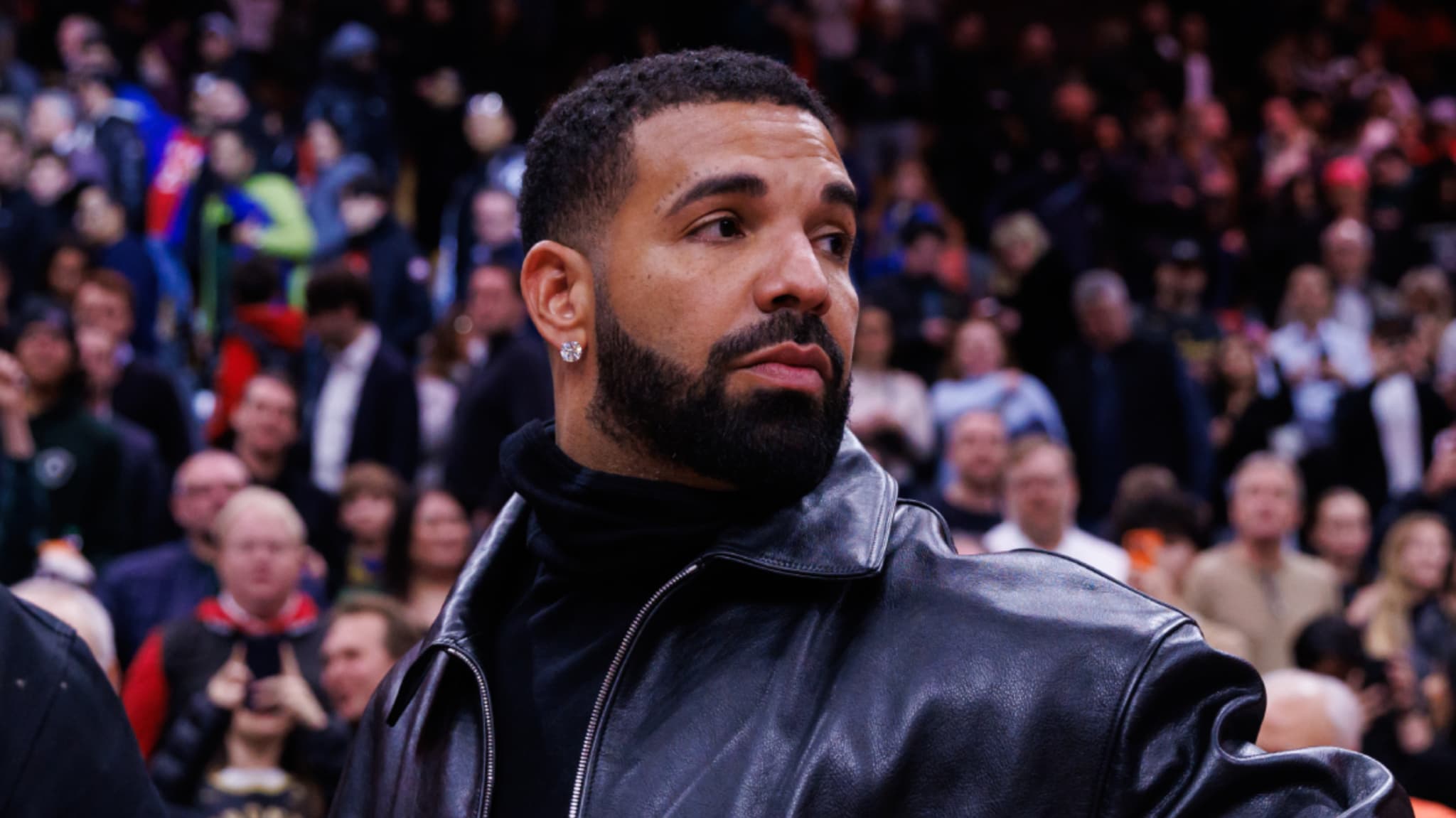 Drake in a leather jacket at a crowded event, looking to the side.