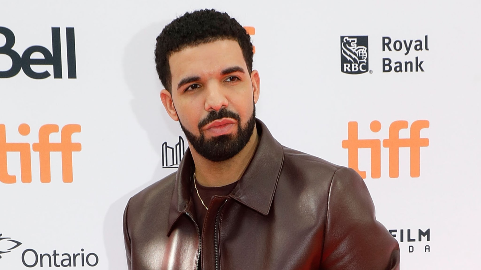 Drake at the Toronto International Film Festival, wearing a brown leather jacket, posing against a backdrop with TIFF logos.
