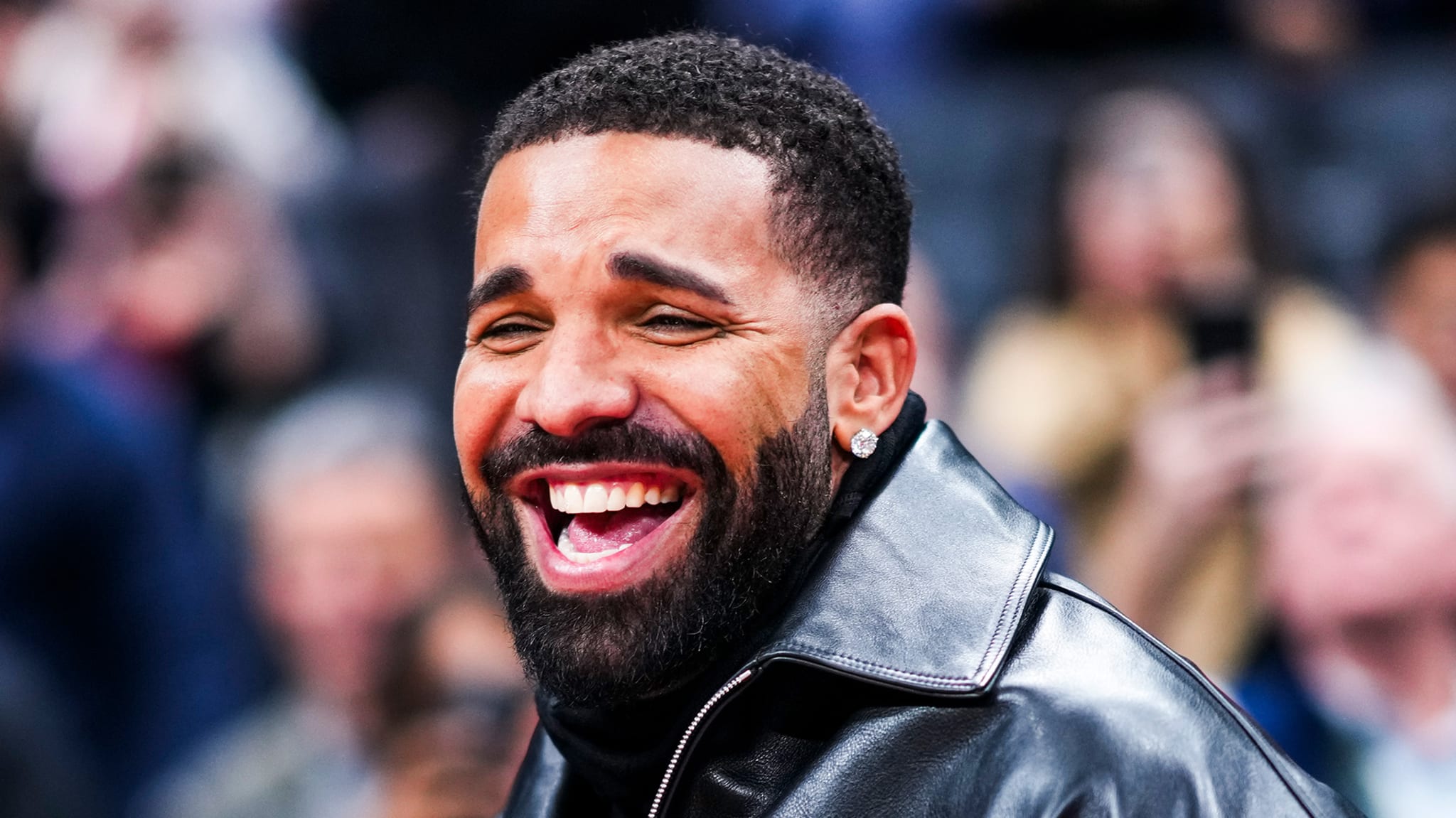 Drake, wearing a black leather jacket, is smiling broadly at an event with a blurred crowd in the background.