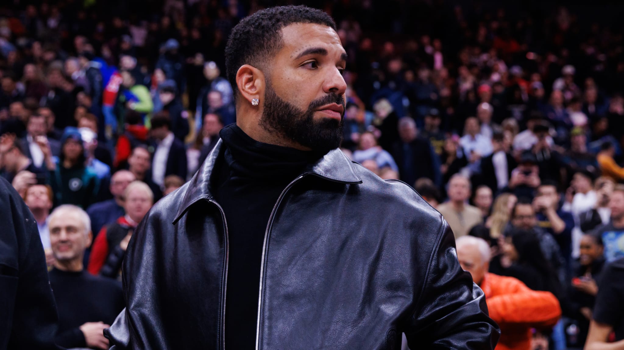 Drake in a black leather jacket at a crowded event, looking to the side.