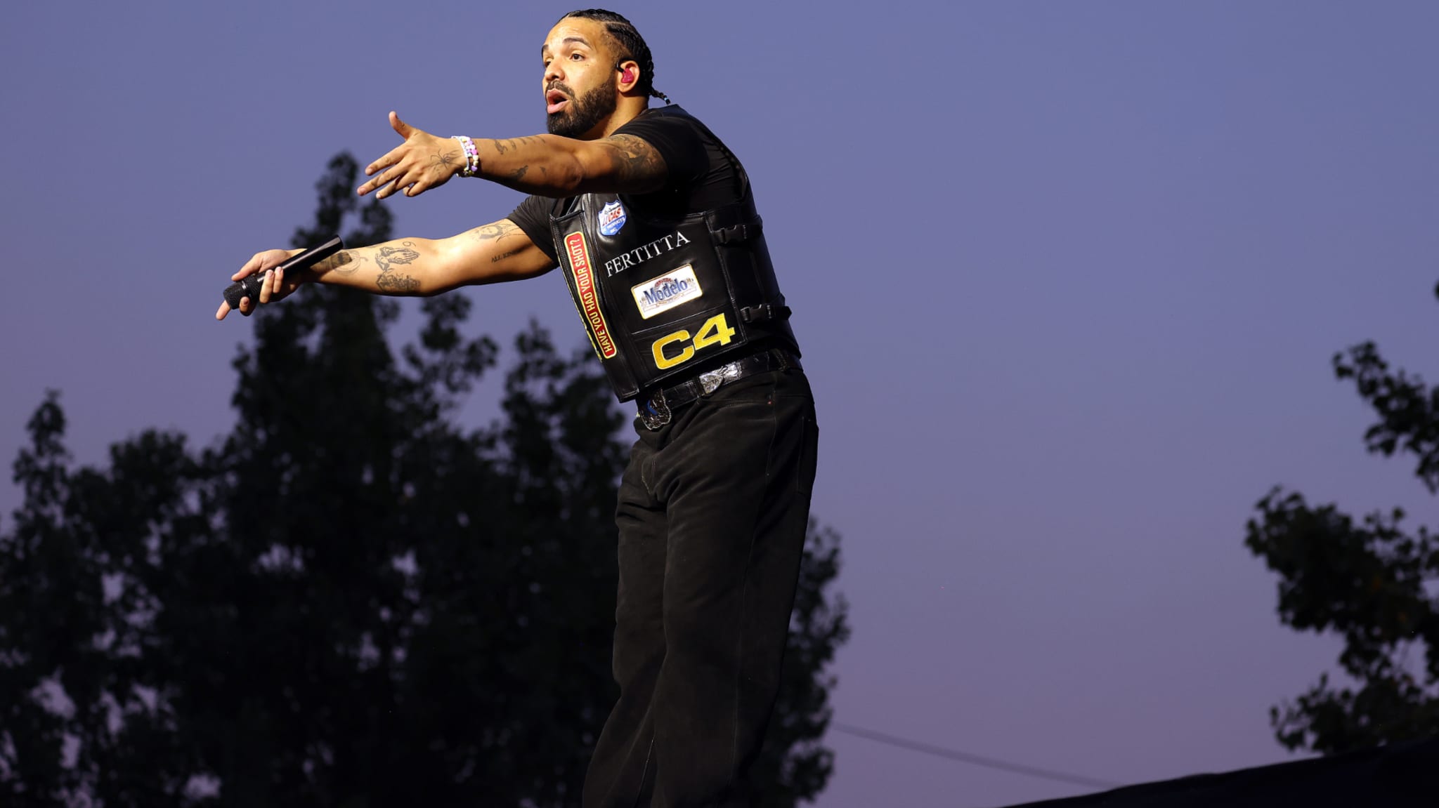 Drake performing on stage, wearing a black outfit with patches, gesturing with his hands, against a twilight sky.