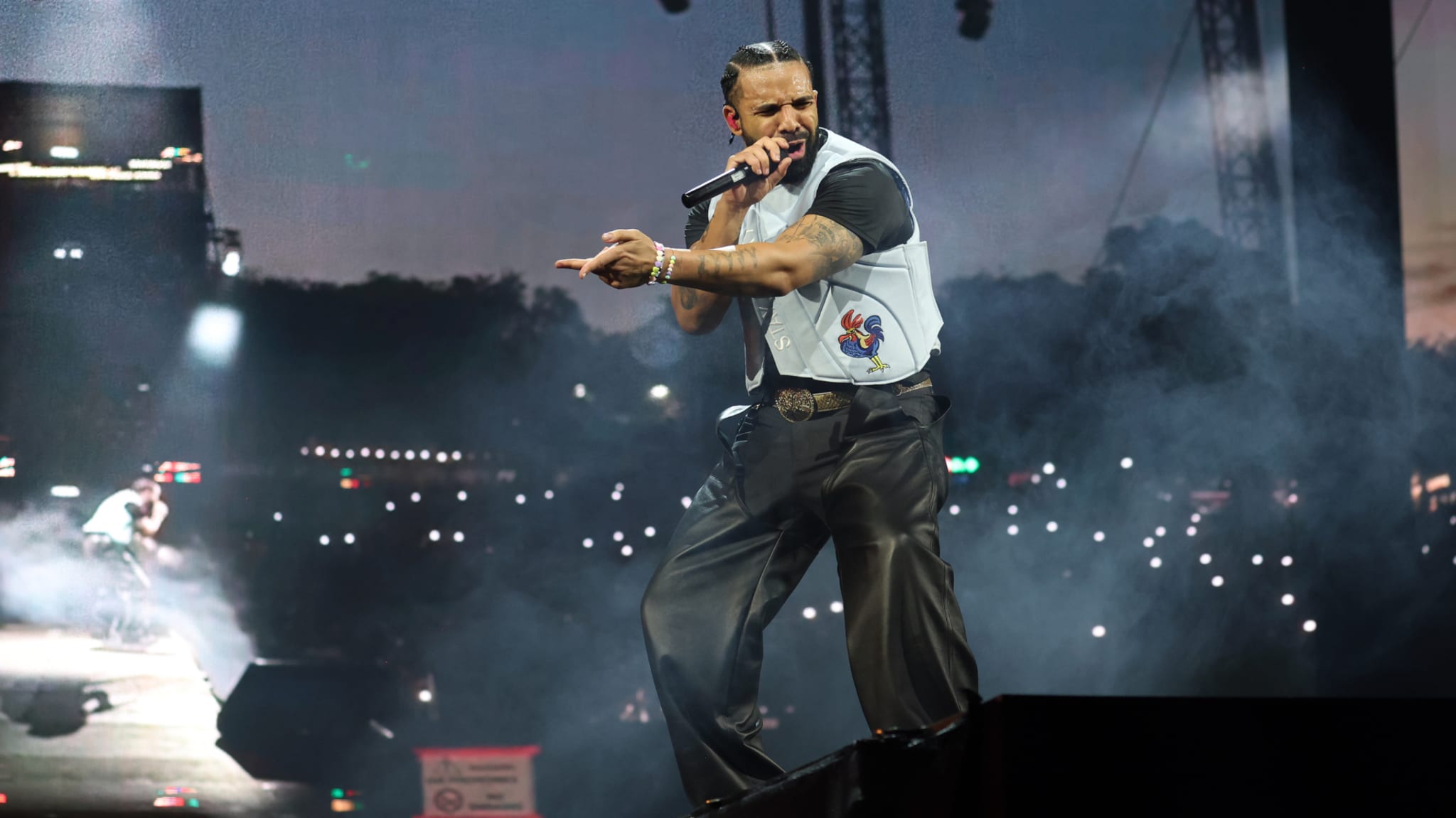 Drake on stage with a microphone, wearing a light vest and dark pants, against a backdrop of a large audience with lights.