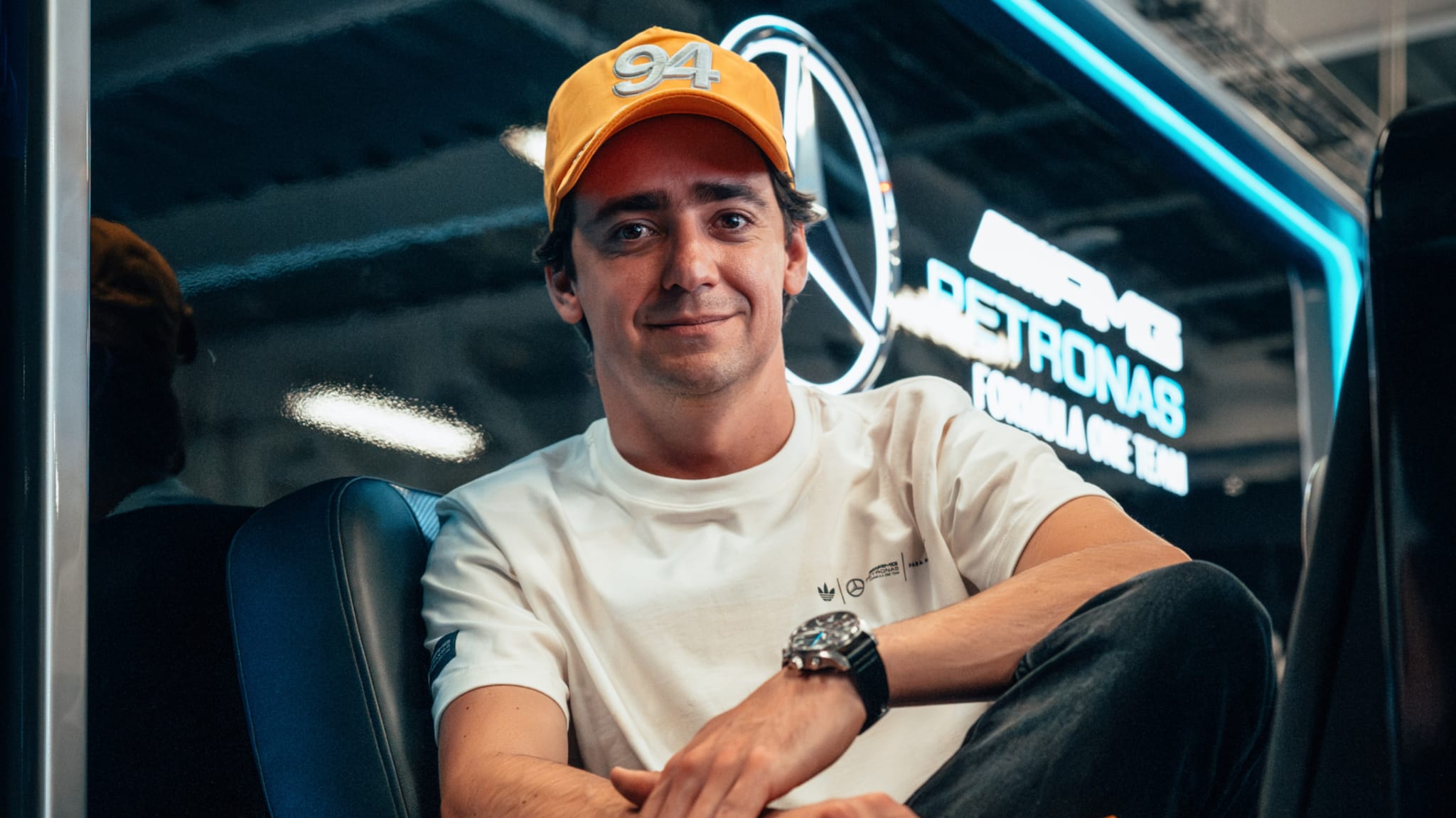 A man wearing a yellow cap with "94" and a white shirt, sitting in front of a Mercedes-AMG Petronas Formula One Team sign.