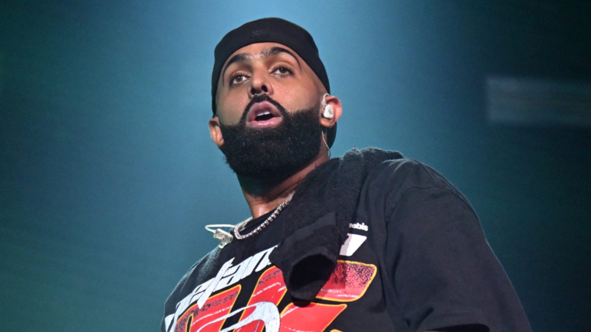 Eladio Carríon with a beard and black cap performing on stage, wearing a graphic t-shirt and a towel around his neck.