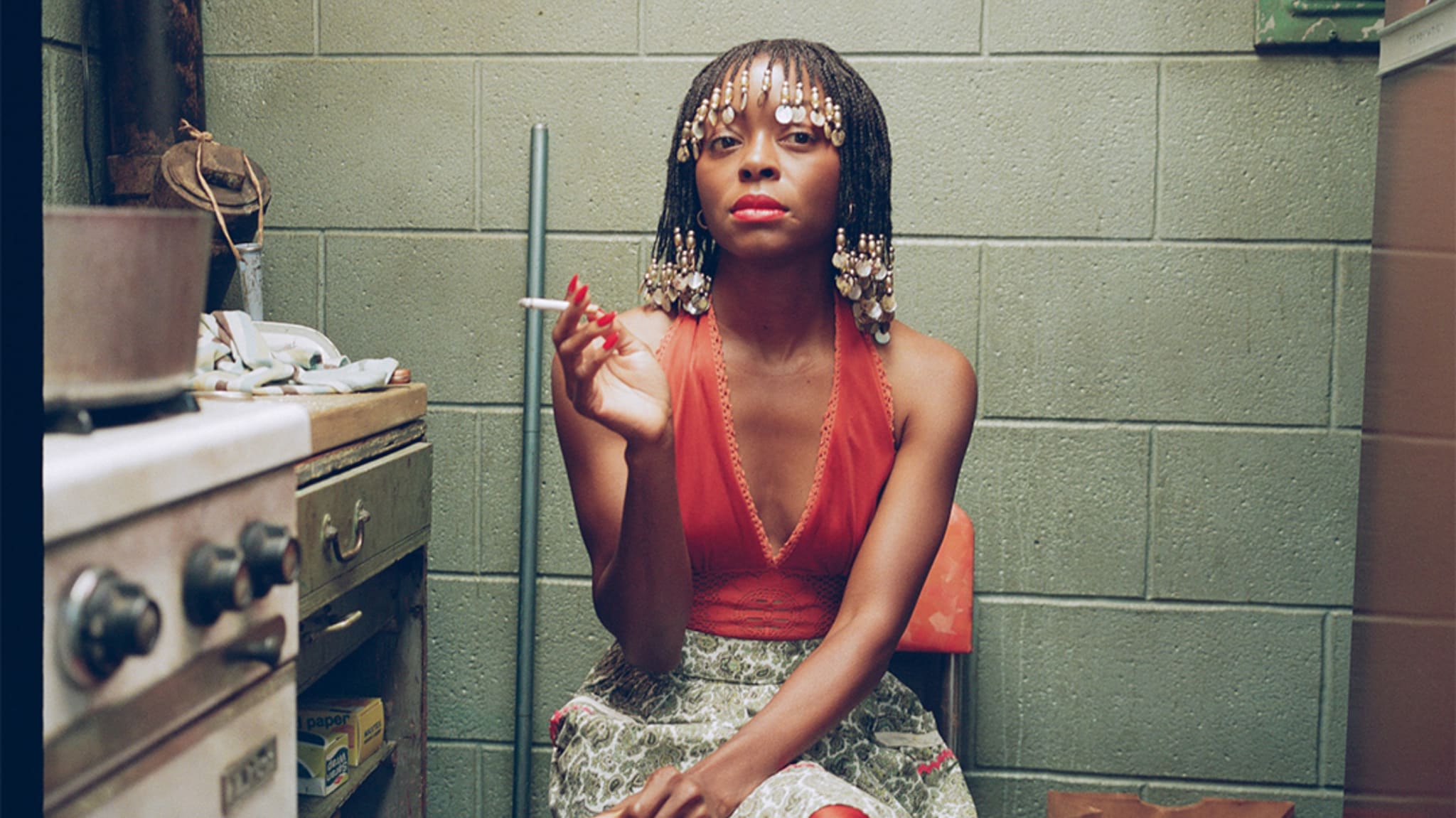 A woman with beaded hair and red lipstick sits in a small kitchen, holding a cigarette. She wears a red top and patterned skirt.