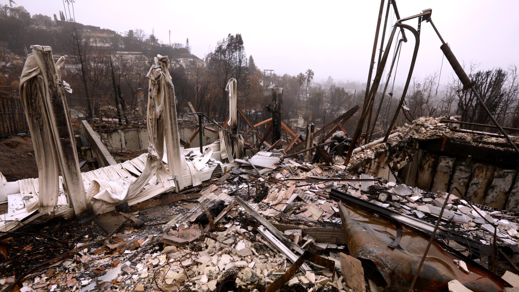 The image shows a landscape of destruction, with charred debris and remnants of structures after a fire, under a gray, overcast sky.