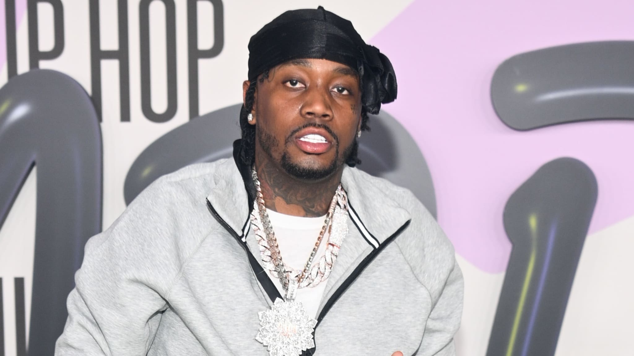 A man wearing a black durag, grey jacket, and large chain necklace stands in front of a backdrop with the words "Hip Hop."