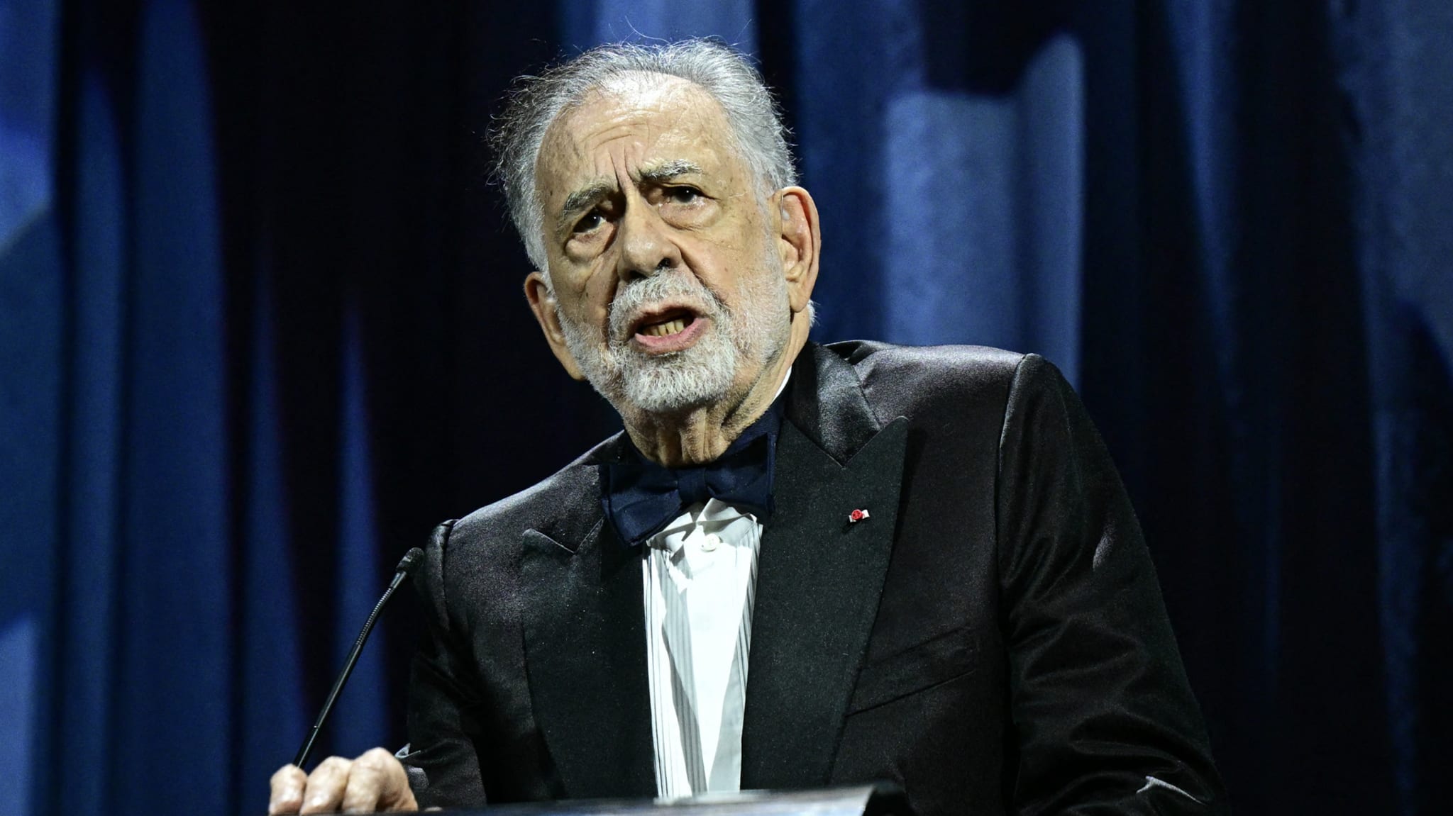 Francis Ford Coppola speaking at an event, wearing a tuxedo with a bow tie, against a dark curtain backdrop.