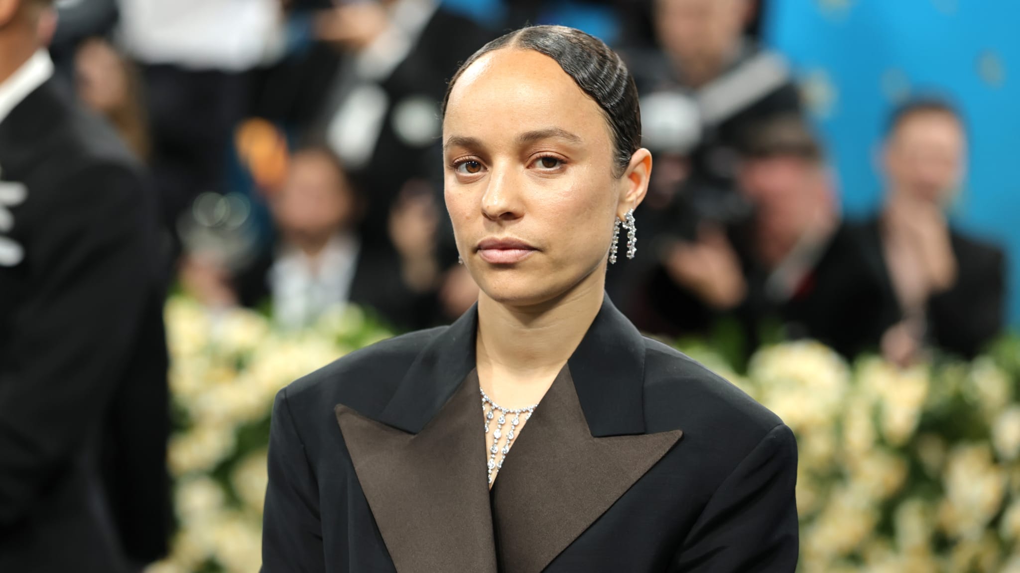 Grace in formal attire with slicked-back hair and earrings stands in front of a blurred crowd at an event.