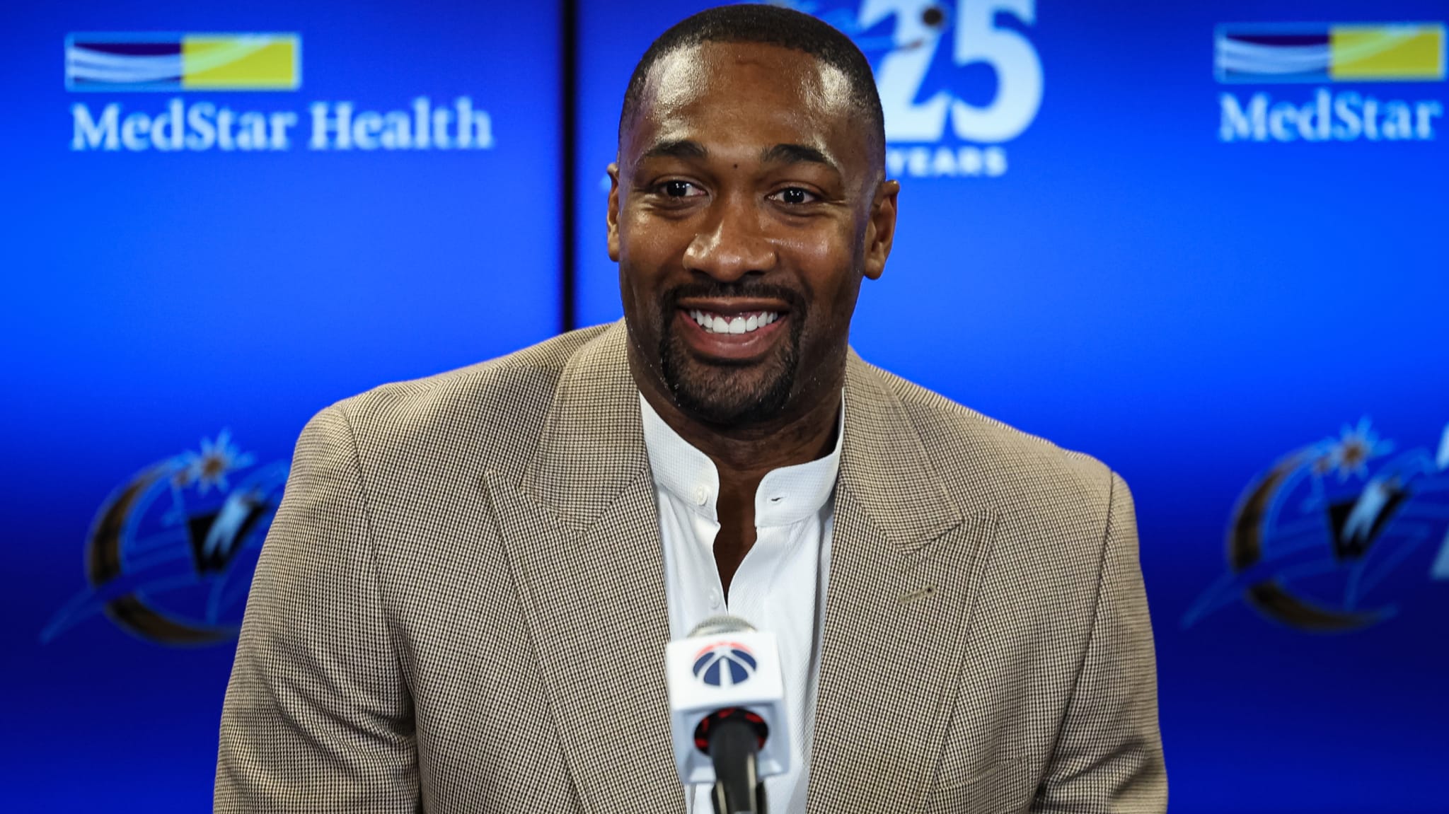 Gilbert Arenas in a beige suit smiles at a press conference, with a blue background featuring "MedStar Health" and a logo.