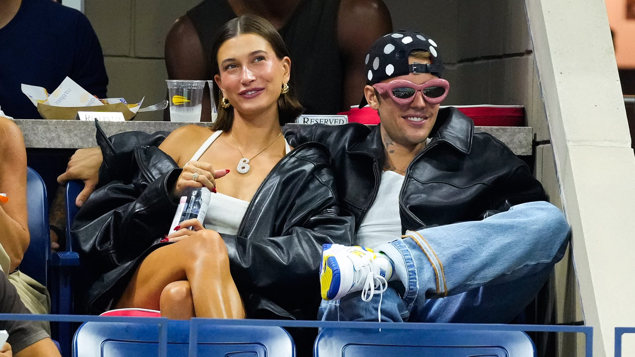 Hailey Bieber and Justin Bieber sitting together at an event, both wearing black jackets. Hailey has a white top, Justin wears a polka dot hat.