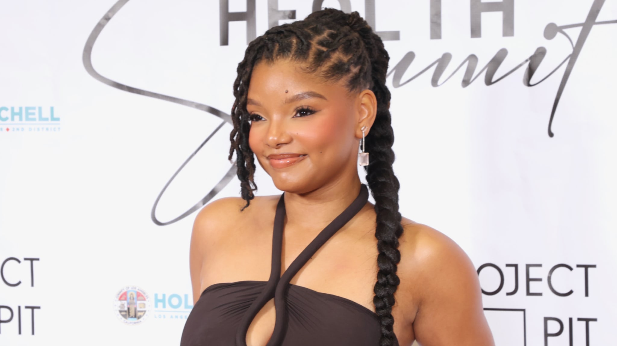 Halle Bailey at an event, wearing a dark halter dress with braided hair. She is smiling against a backdrop with text.