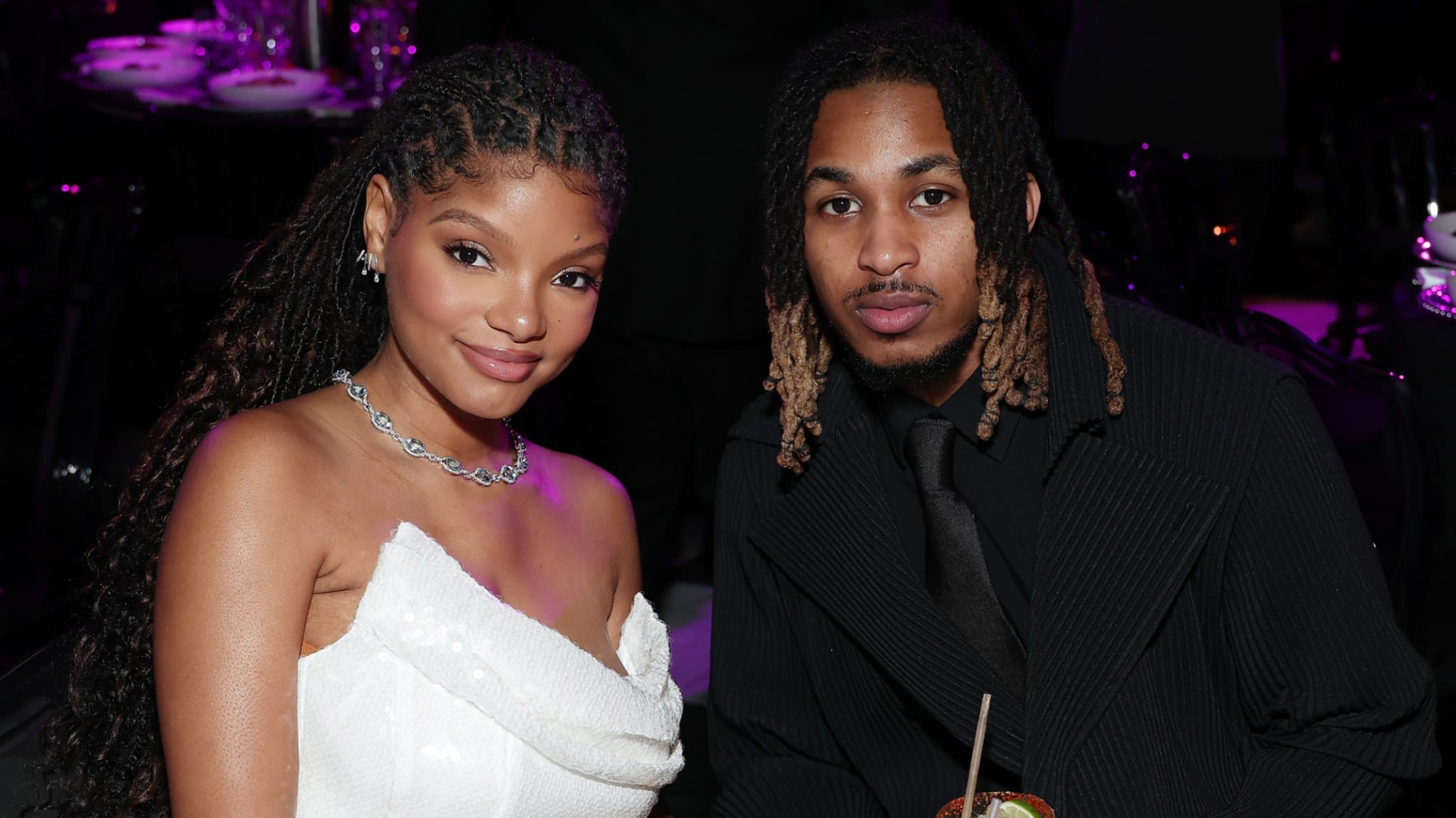 Halle Bailey in a white dress and DDG in a black suit pose together at an event with a dimly lit, purple background.