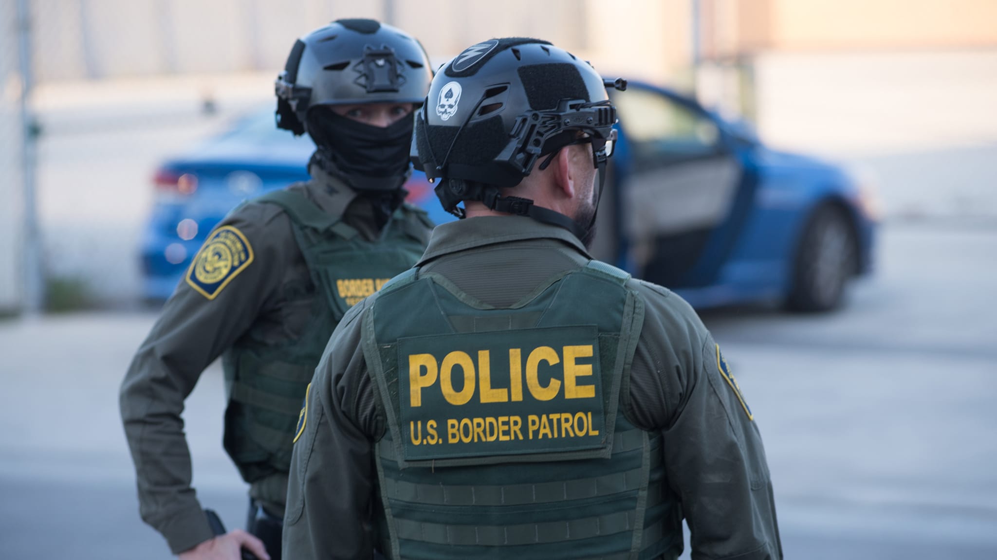 Two U.S. Border Patrol officers in tactical gear and helmets stand near a blue car.