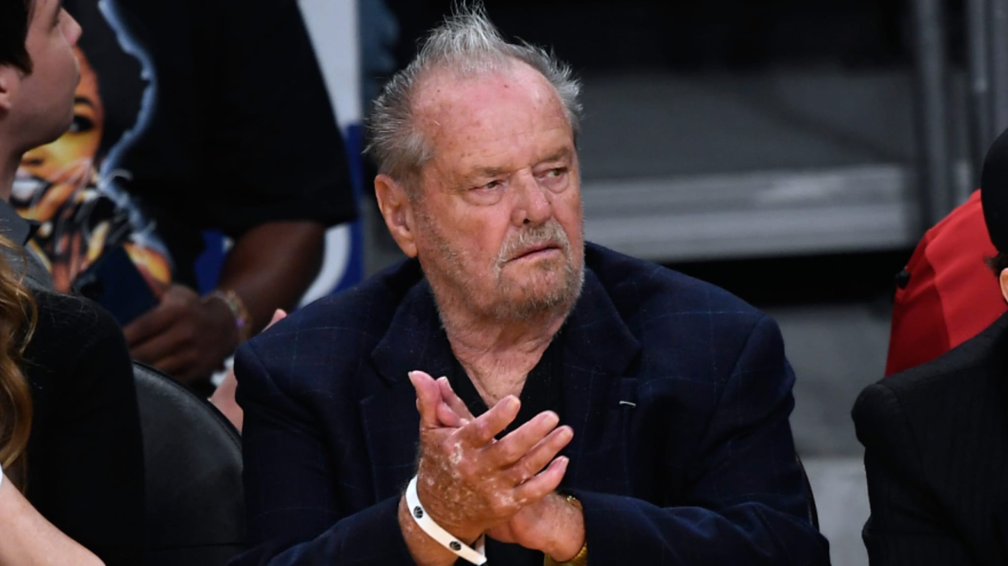 Jack Nicholson with a beard clapping at a sports event, wearing a dark suit jacket.