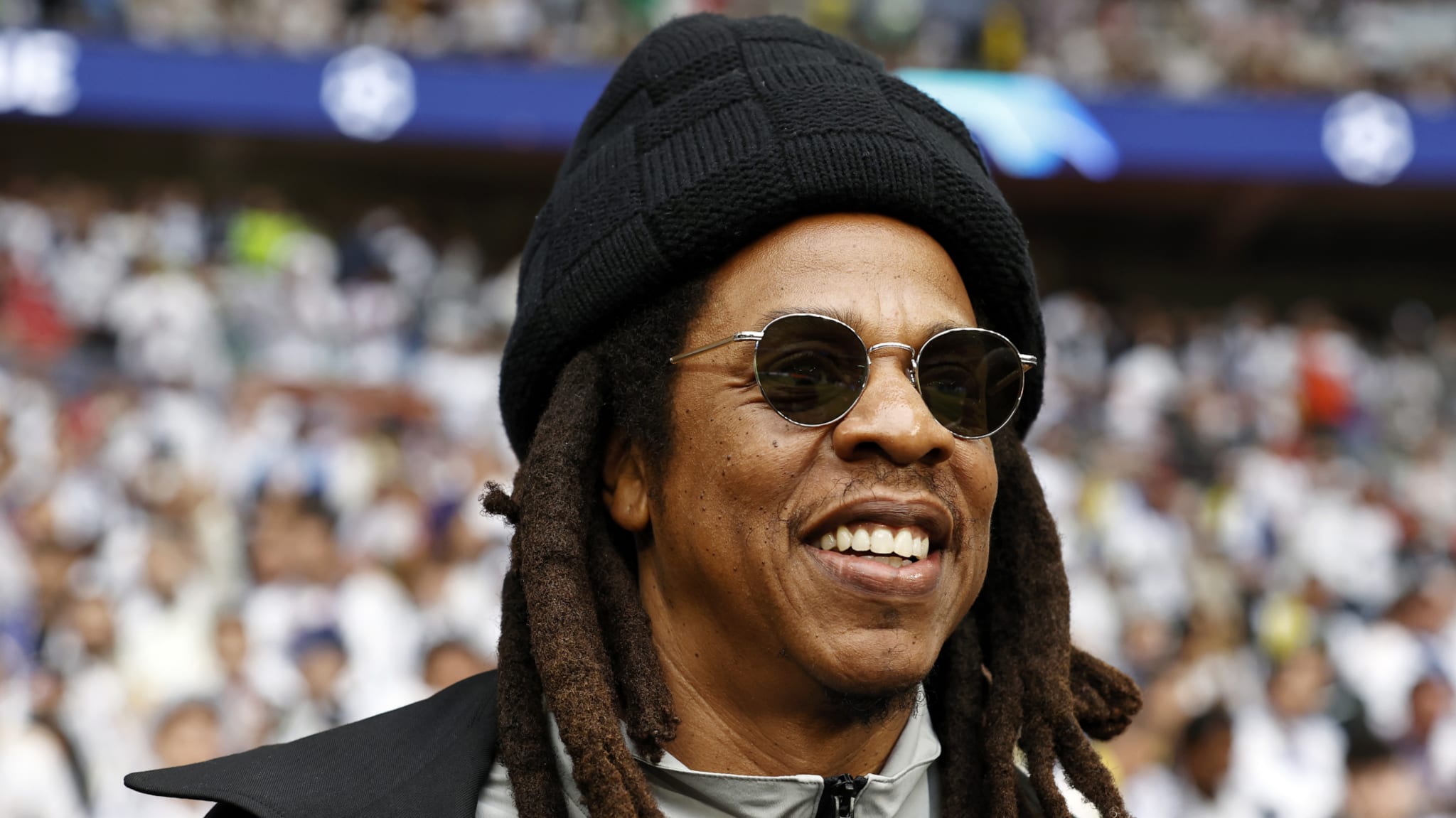 Jay-Z wearing sunglasses and a beanie, smiling at an event with a crowd in the background.