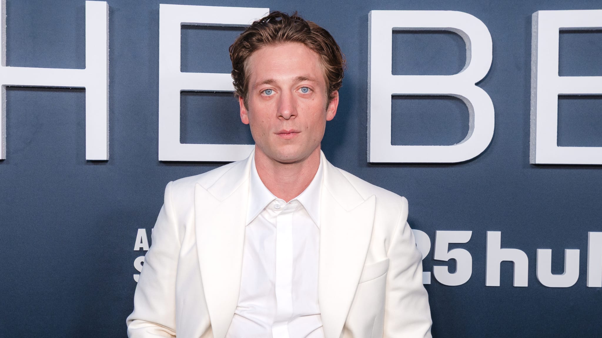A man in a white suit poses in front of a backdrop with text.