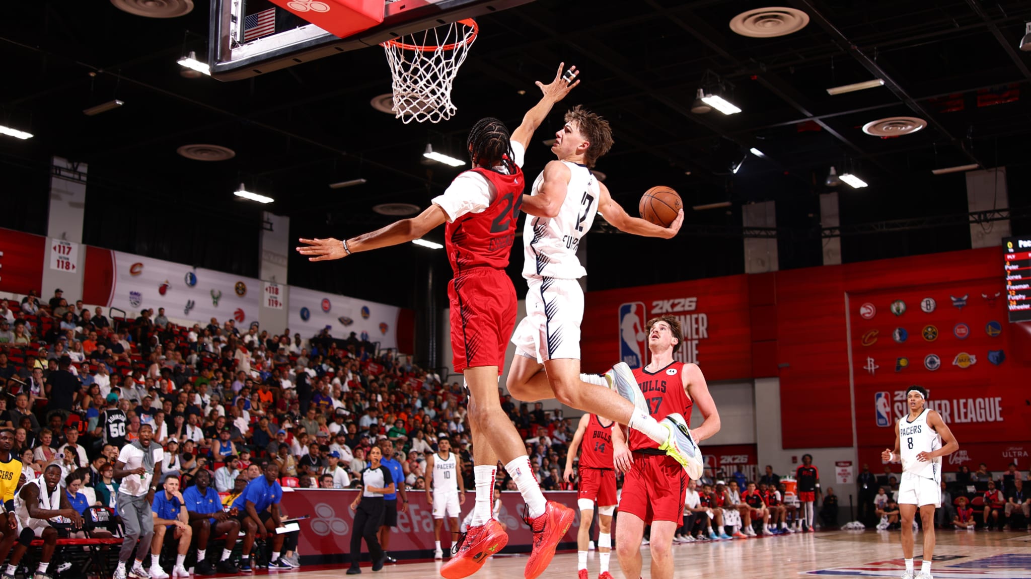 Two basketball players in mid-air near the hoop, one in a white jersey and the other in red, during a game with a cheering crowd.
