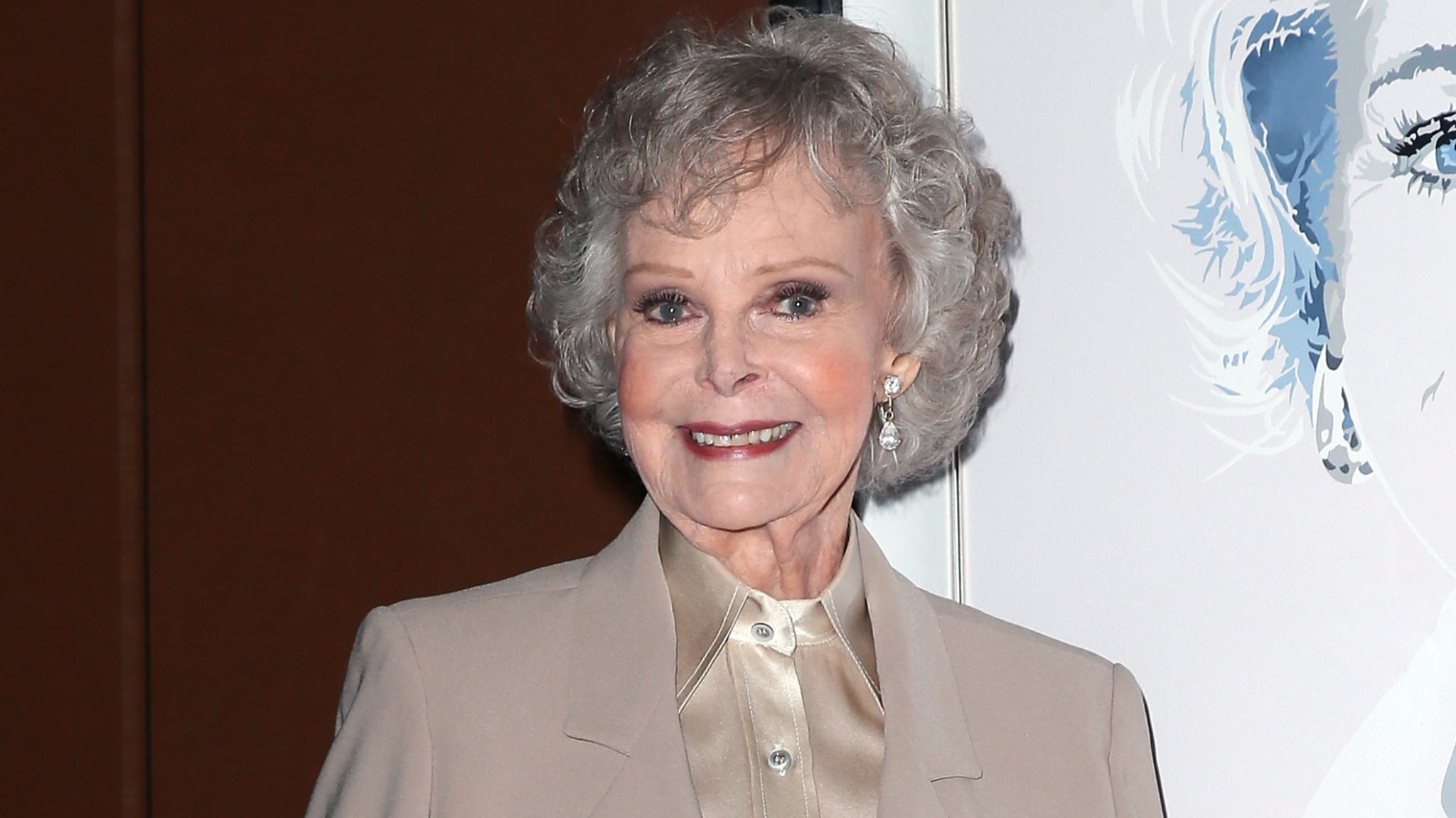 An older woman with curly gray hair and a beige suit smiles in front of a stylized portrait.