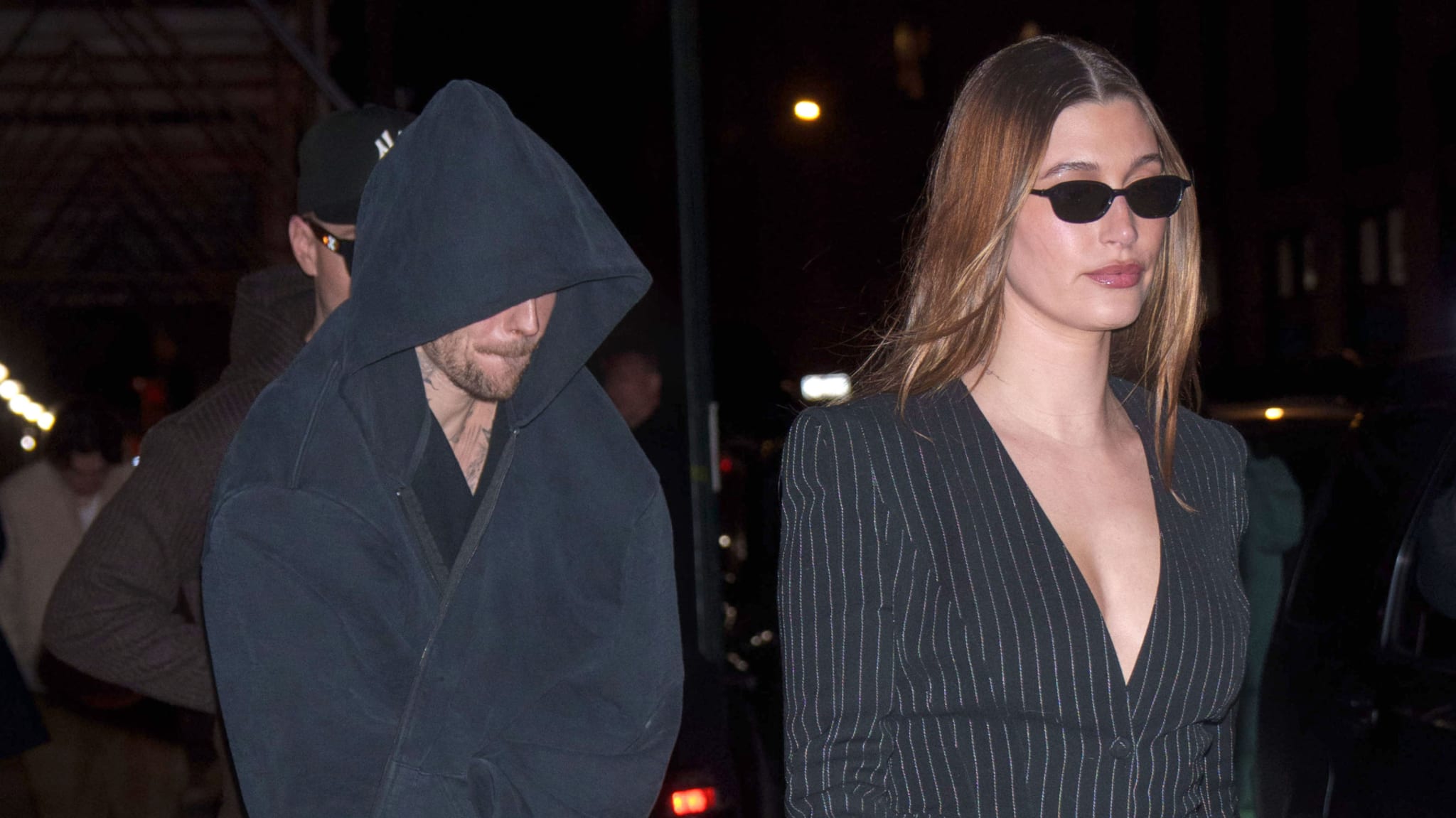 Justin Bieber in a hooded jacket walks beside Hailey Bieber, who is wearing sunglasses and a pinstripe outfit, at night.
