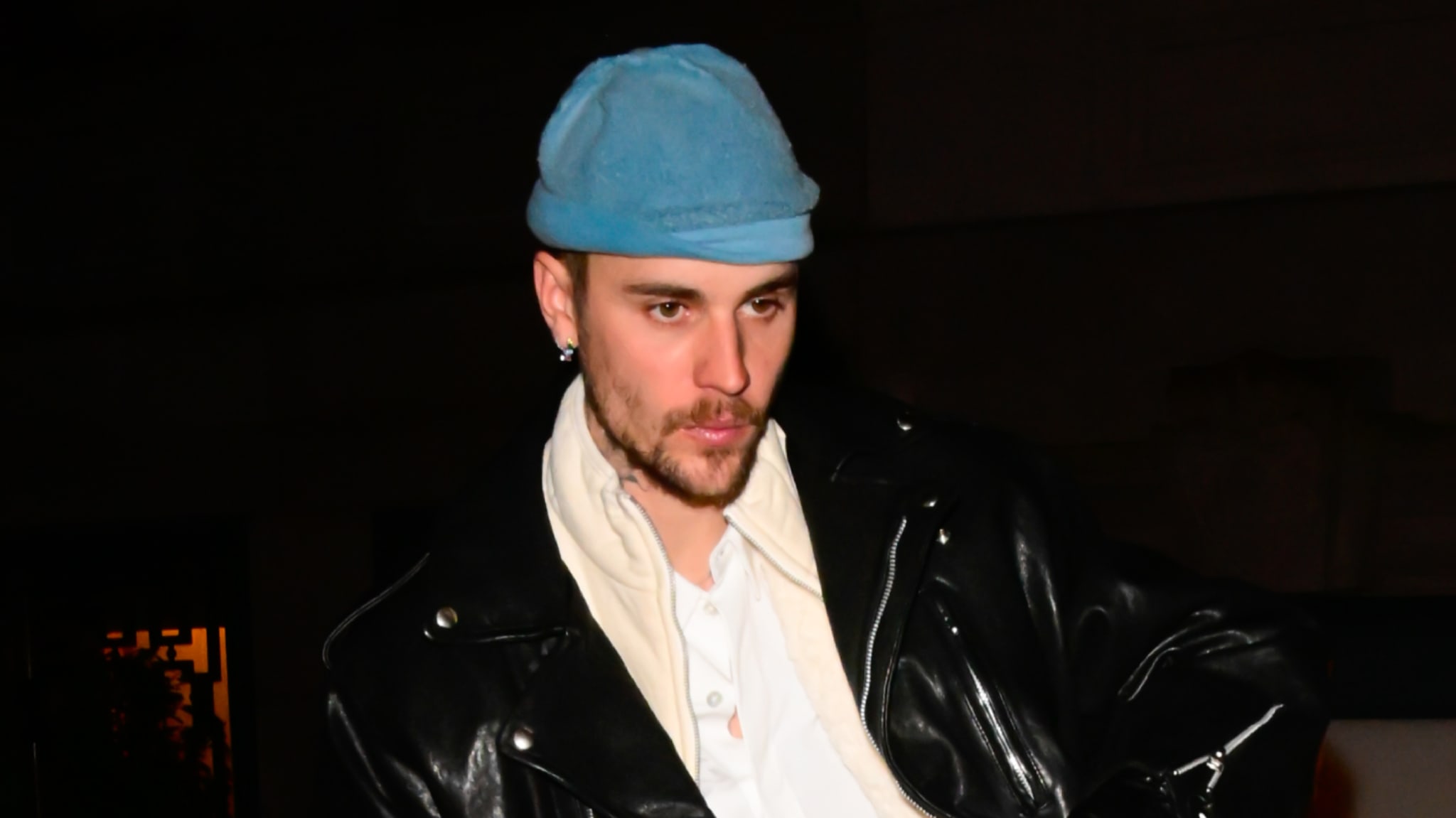 Justin Bieber wearing a blue beanie, black leather jacket, and white shirt, standing outdoors at night.
