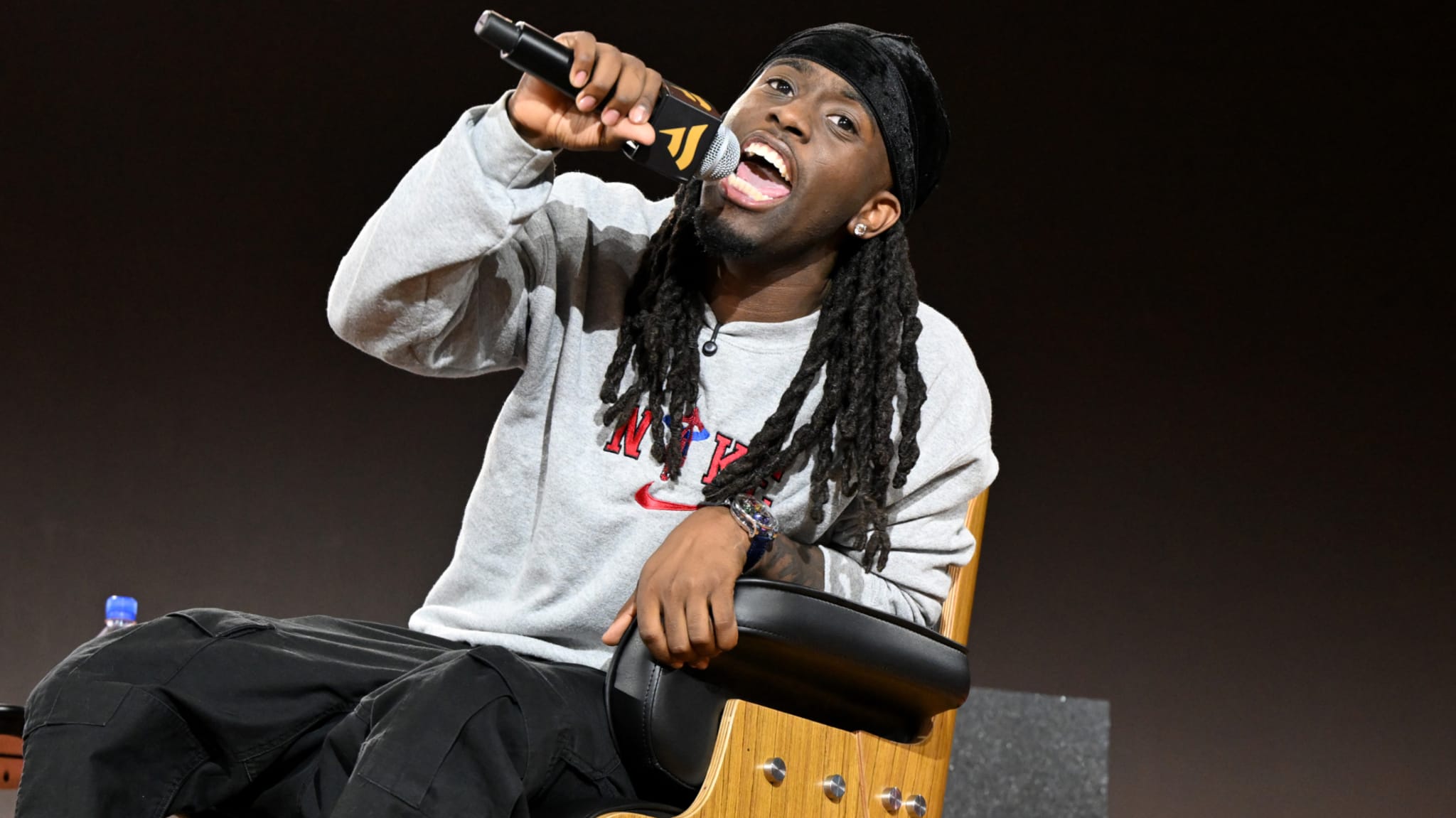 Kai Cenat with long hair and a hat is sitting in a chair, holding a microphone, and speaking enthusiastically.
