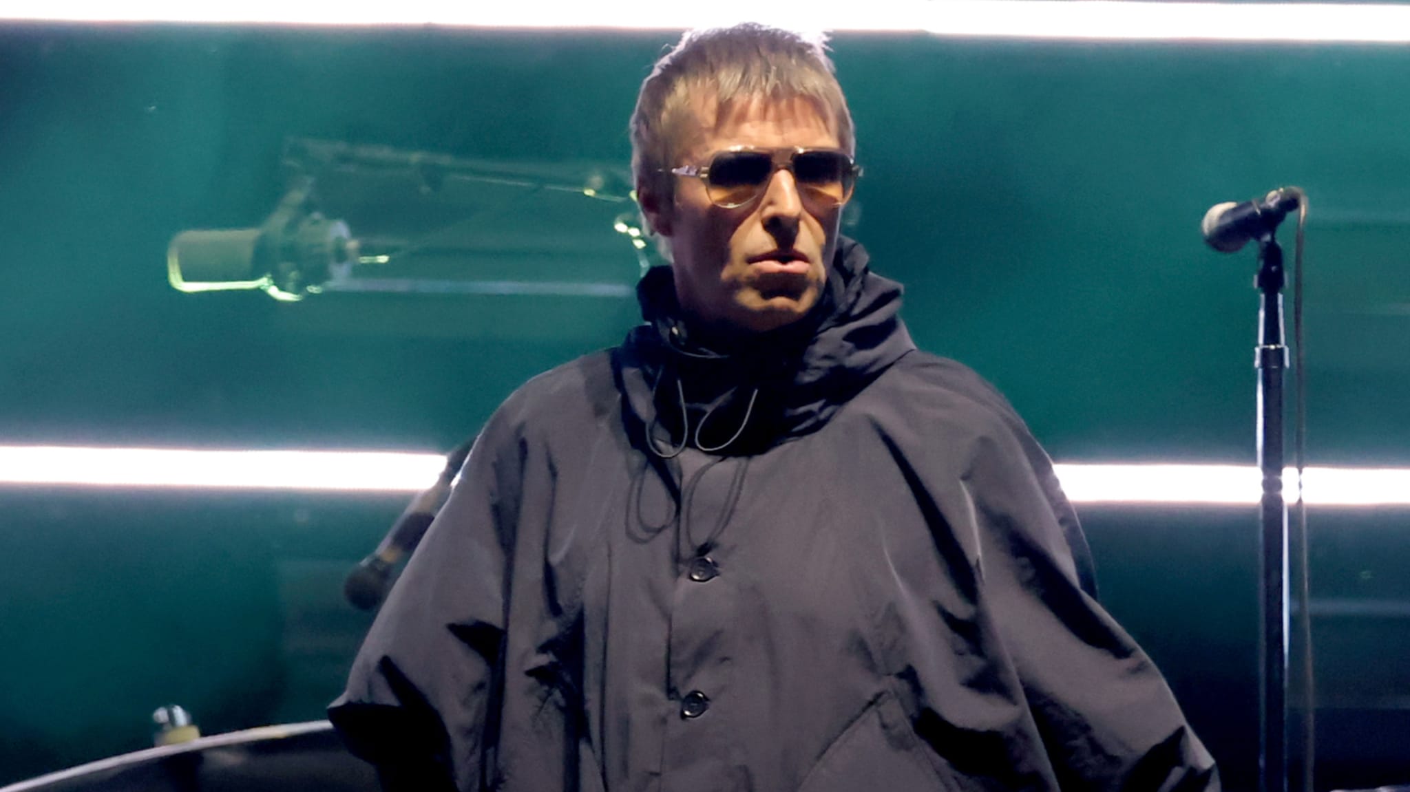 A man in sunglasses and a dark jacket stands on stage with a microphone, under bright horizontal lights.