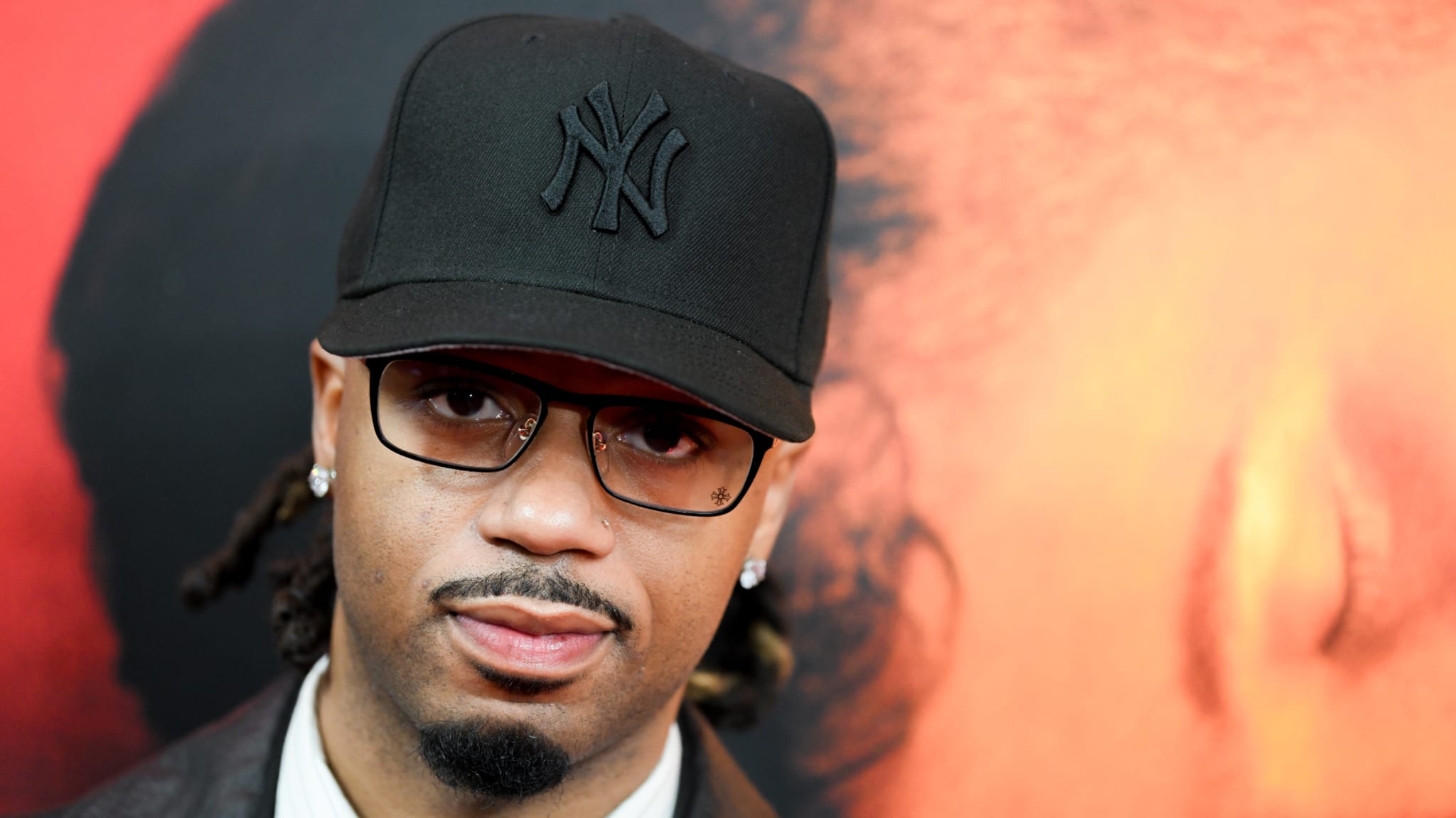 Metro Boomin wearing glasses and a black New York Yankees cap, with a blurred background featuring warm colors.