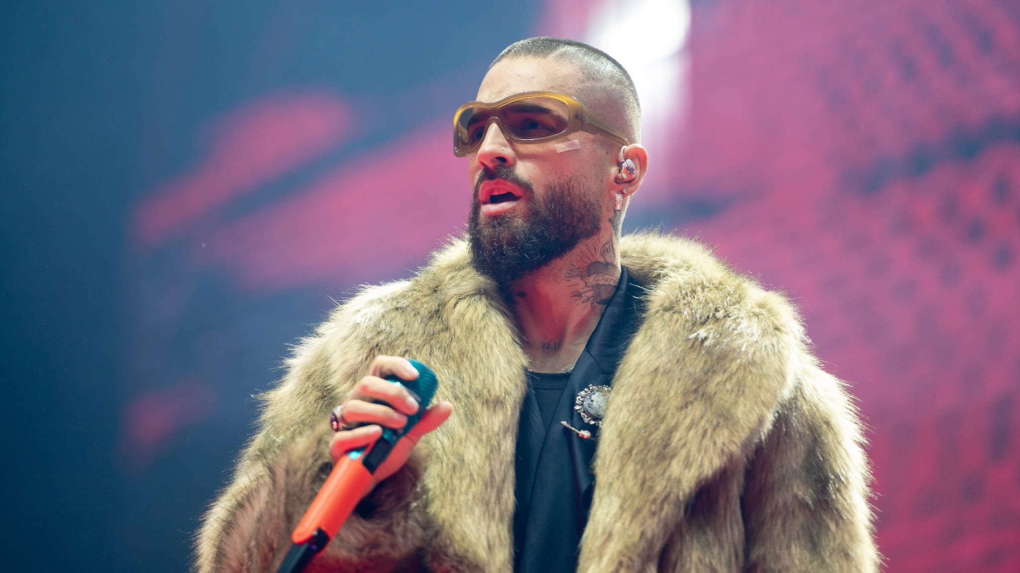 Colombian singer Maluma with sunglasses and a fur coat holds a microphone on stage, with a red background.