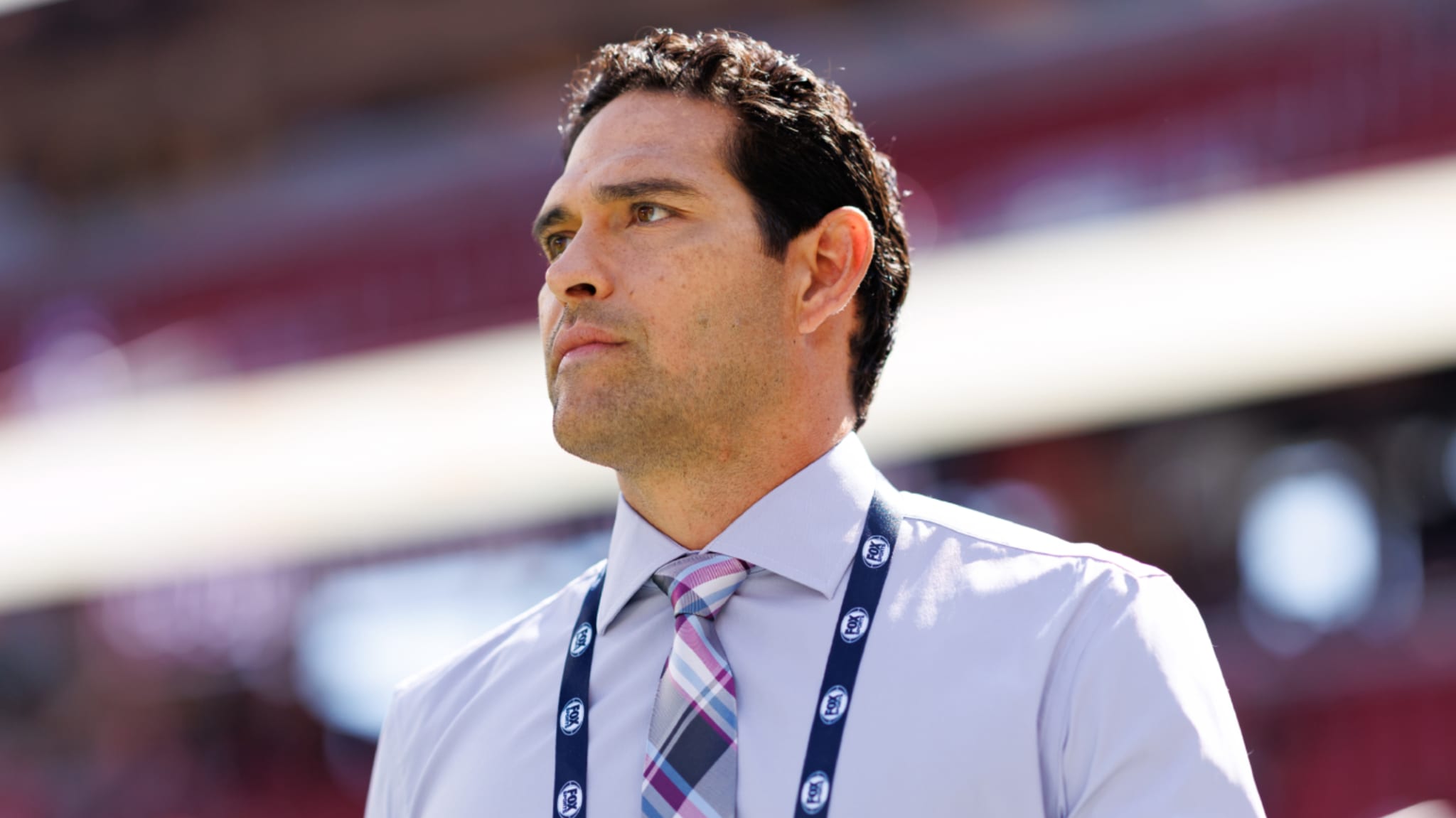 Mark Sanchez in a dress shirt and tie, wearing a lanyard, stands outdoors with a focused expression.