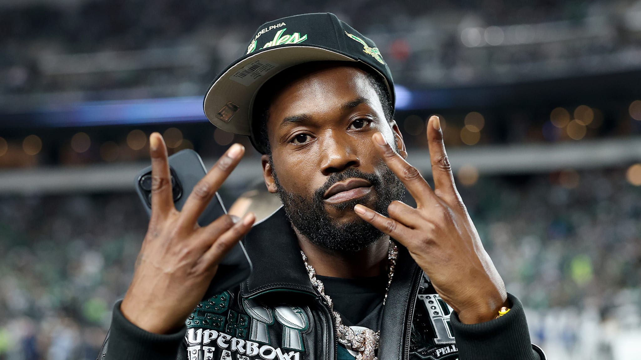 Rapper Meek Mill wearing a Philadelphia Eagles cap and jacket, making a hand gesture at a sports event.