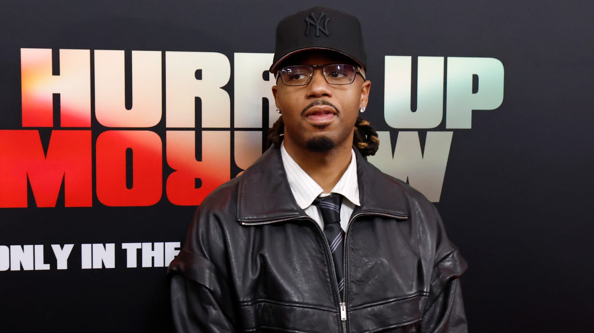 Metro Boomin wearing a black cap, glasses, leather jacket, and tie stands in front of a backdrop with text.