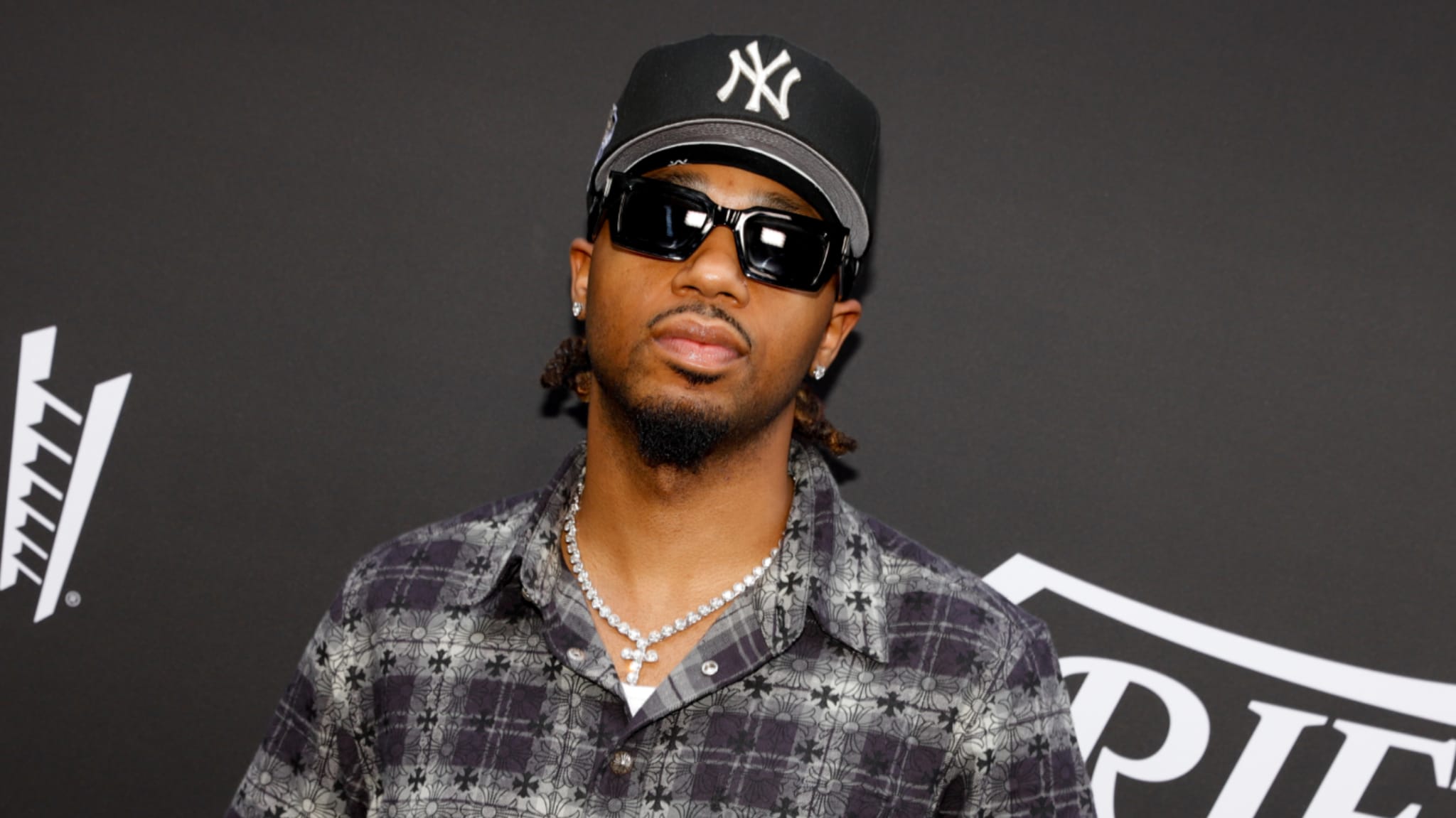 Metro Boomin wearing a New York Yankees cap, sunglasses, and a patterned shirt, standing against a dark backdrop.