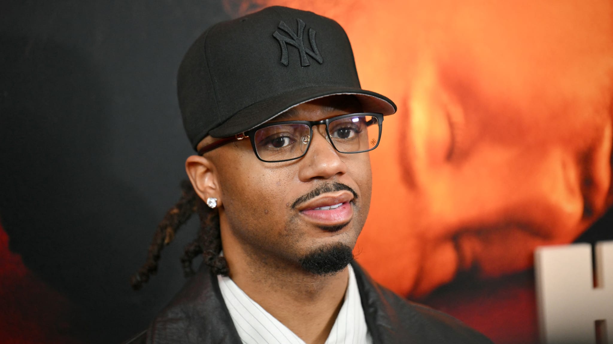 Metro Boomin wearing a black New York Yankees cap, glasses, and a leather jacket stands in front of a red and orange background.