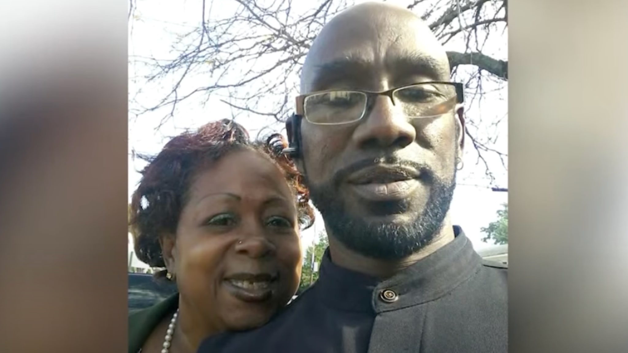 A man and woman pose for a selfie outdoors. The man wears glasses and a Bluetooth earpiece, while the woman smiles in the background.