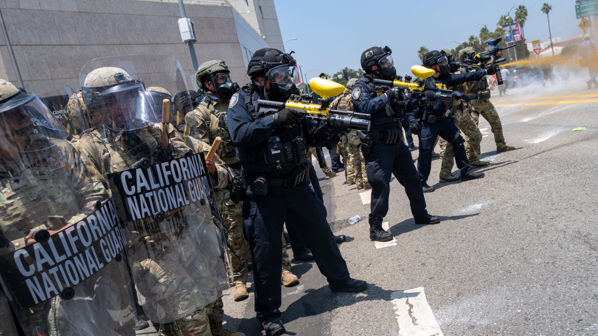 Police and California National Guard in riot gear and uniforms, holding shields and paintball guns, stand in formation on a street.