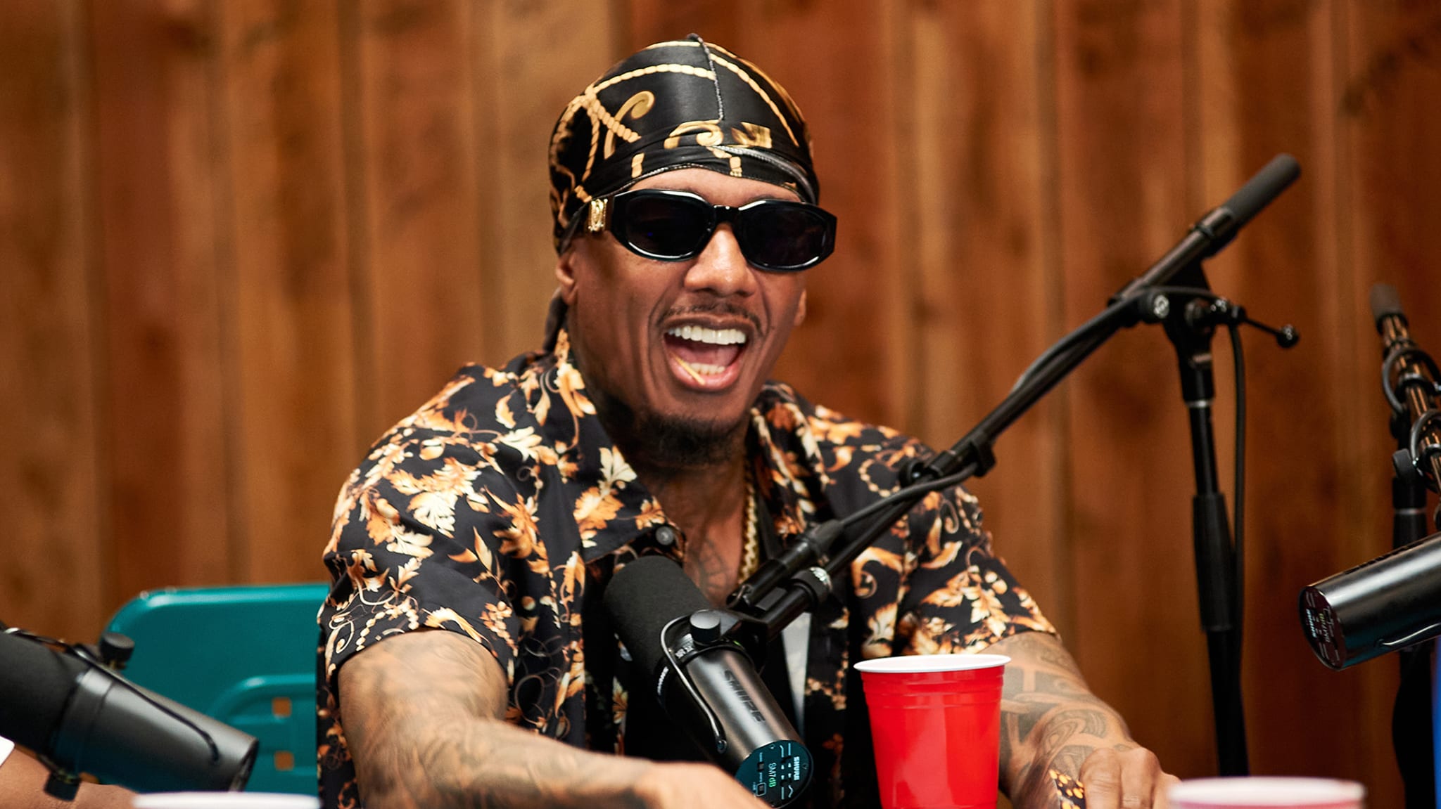 Nick Cannon wearing sunglasses and a patterned shirt, speaking into a microphone, smiling, with a red cup on the table.