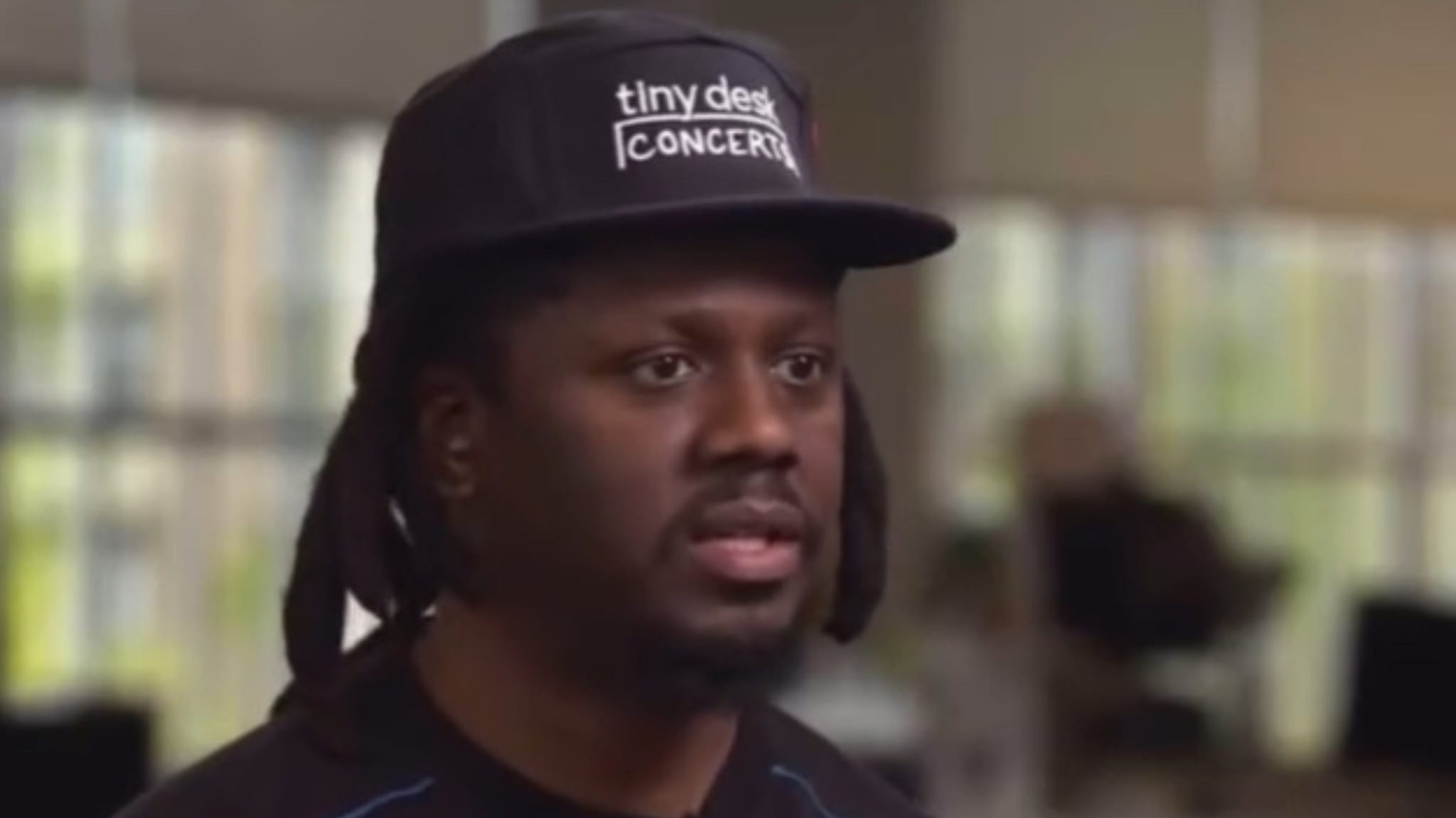 A person wearing a "Tiny Desk Concerts" cap is speaking indoors, with a blurred background.