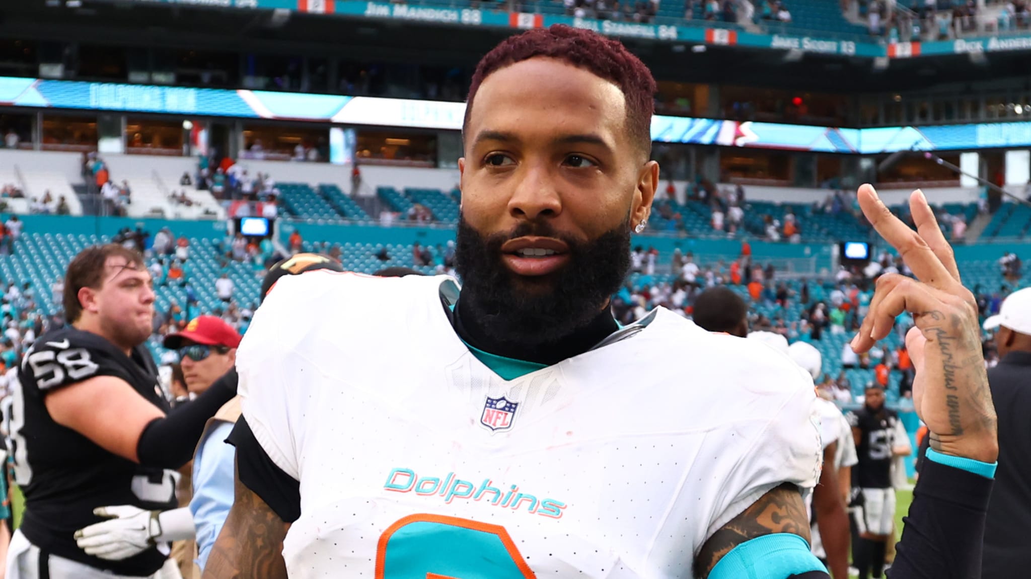 A football player in a Miami Dolphins jersey on the field, gesturing with a peace sign. The stadium and other players are in the background.