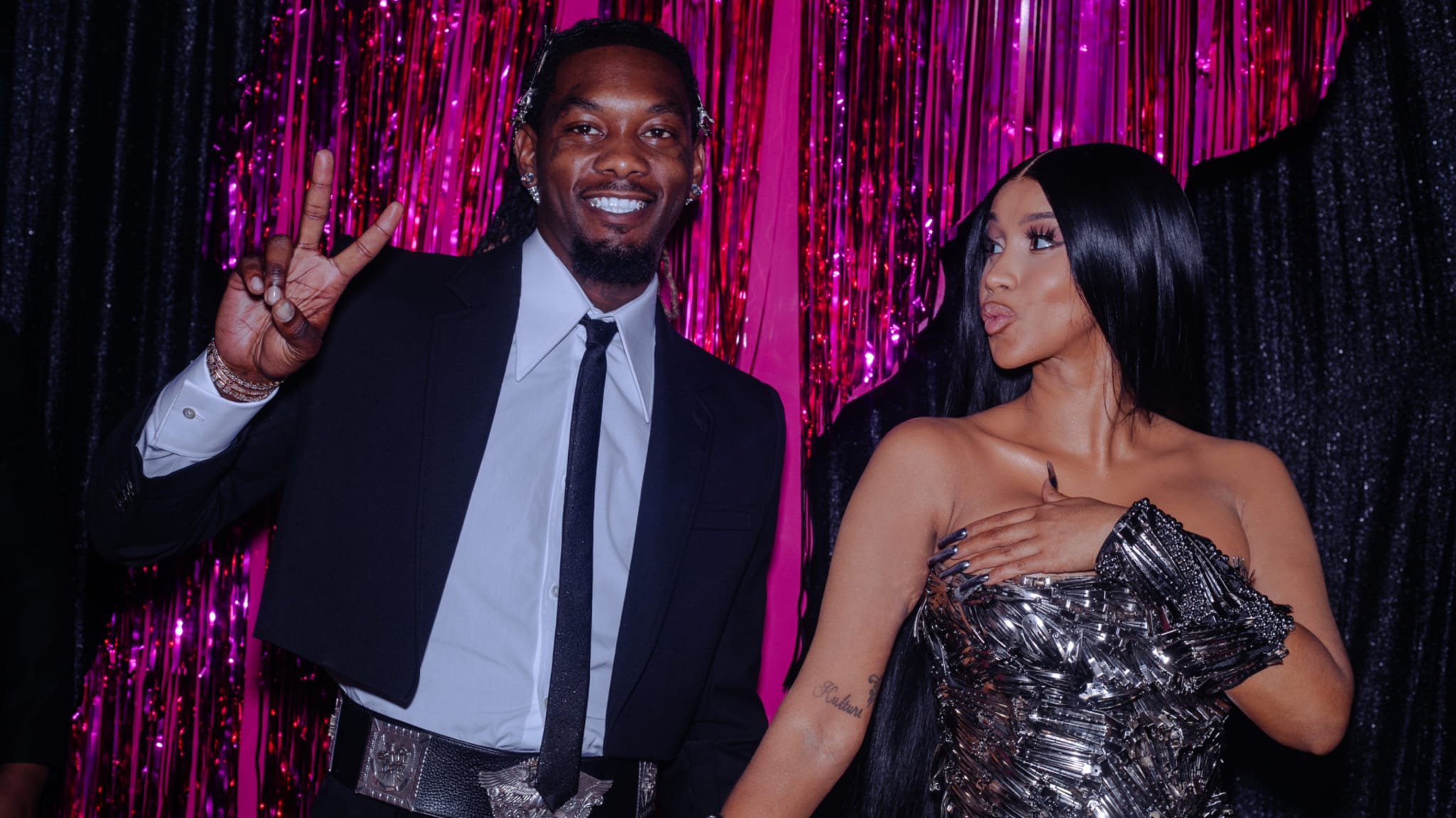 Offset and Cardi B pose together. Offset wears a suit and flashes a peace sign, while Cardi B is in a metallic dress, set against pink tinsel.