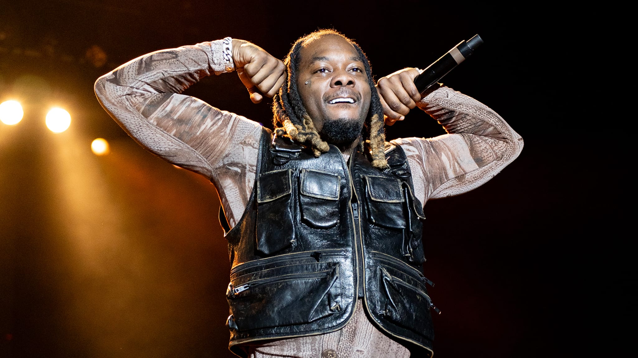 Offset performing on stage, wearing a black vest and holding a microphone, with bright stage lights in the background.