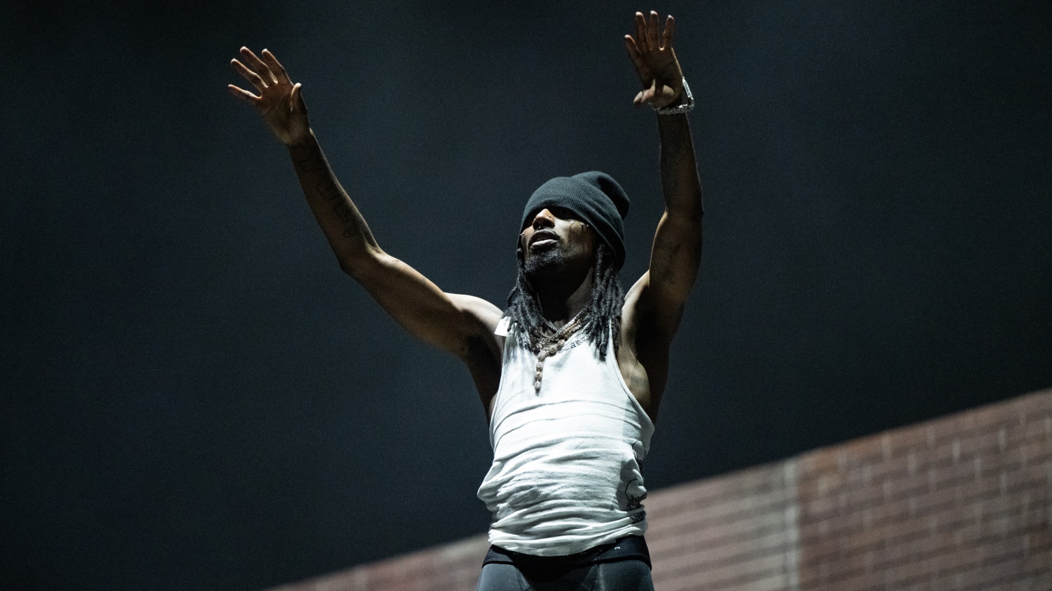 Playboi Carti wearing a beanie and tank top raises their arms on stage, with a dark background and a brick wall behind them.