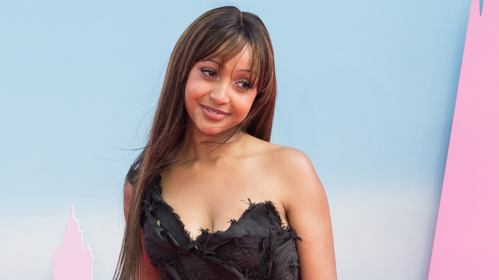 PinkPantheress in a black dress poses against a pastel background, smiling slightly with long hair framing her face.