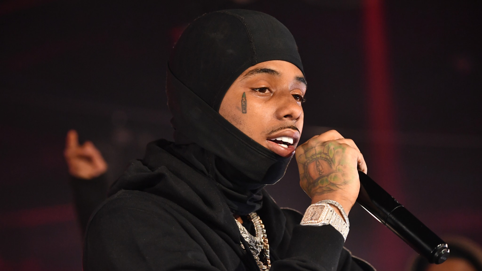 Pooh Shiesty wearing a black hooded outfit, performing on stage with a microphone. They have tattoos on their face and hand.