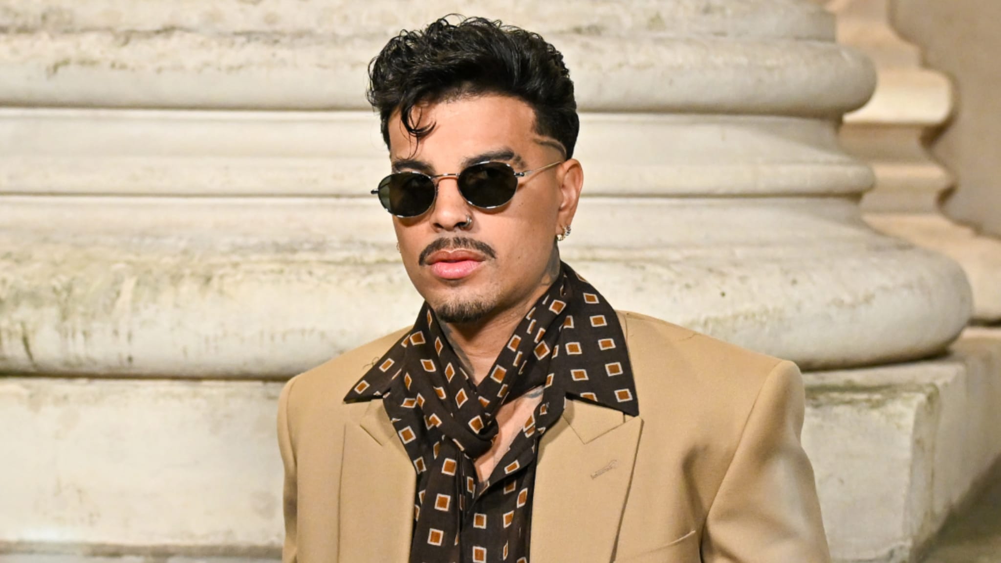 Rauw Alejandro wearing sunglasses and a stylish tan suit with a patterned shirt and scarf, standing in front of a stone pillar.