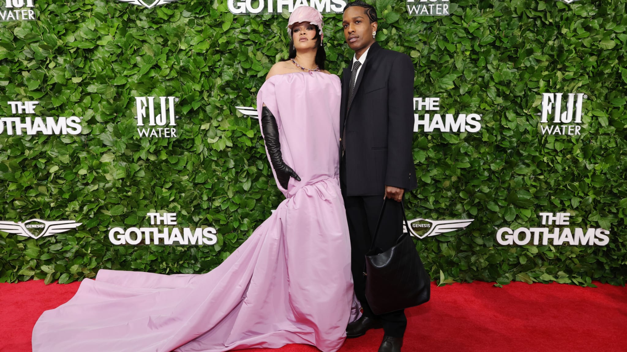 Rihanna in a flowing pink gown and A$AP Rocky in a black suit pose on the red carpet at The Gothams, with a leafy backdrop.